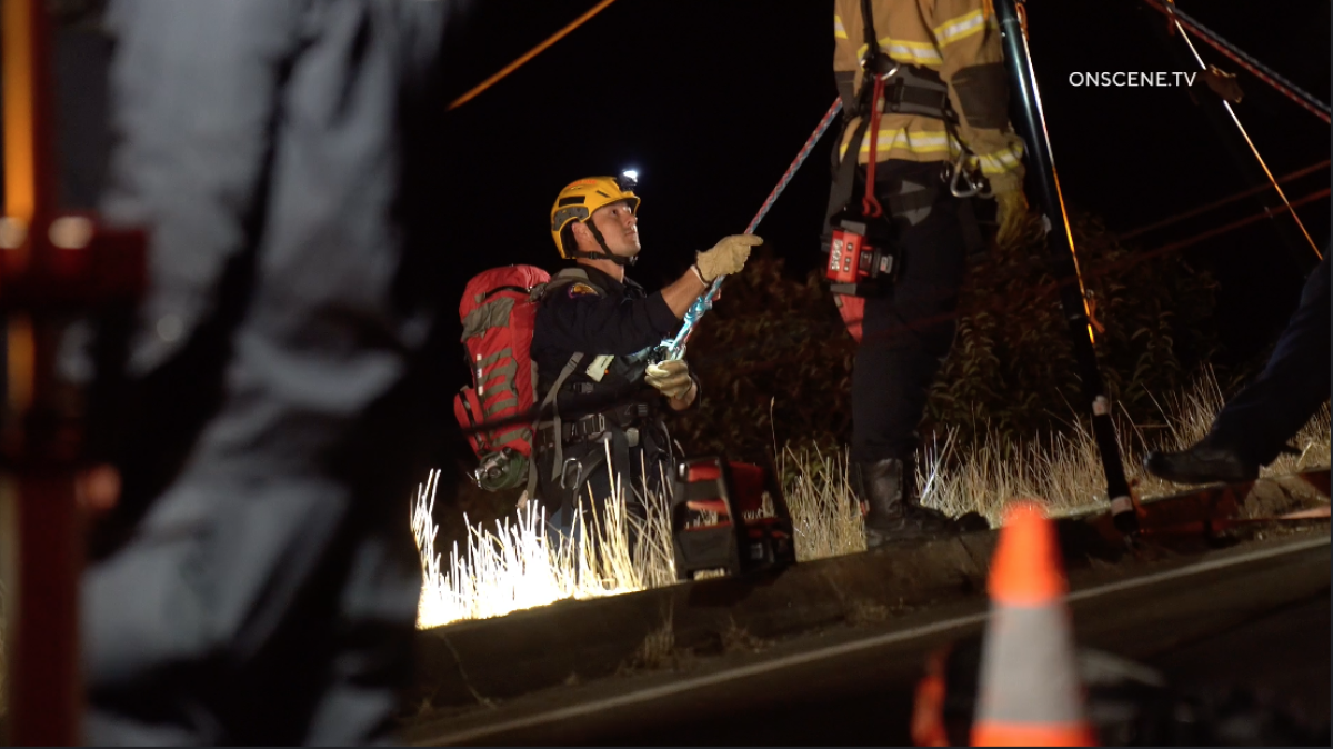 A man in a helmet with a light and a florescent backpack rappels down the side of a cliff in the dark.