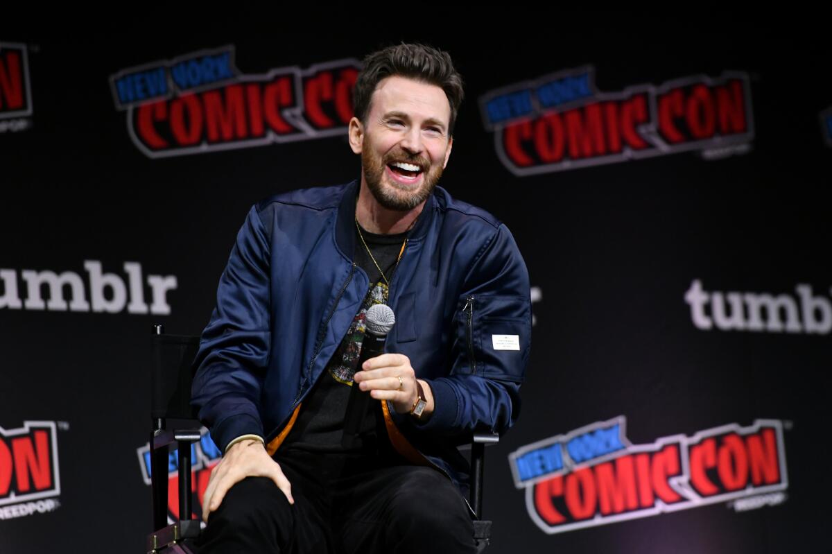 Chris Evans holds a microphone while sitting in a chair onstage in front of a New York Comic Con backdrop