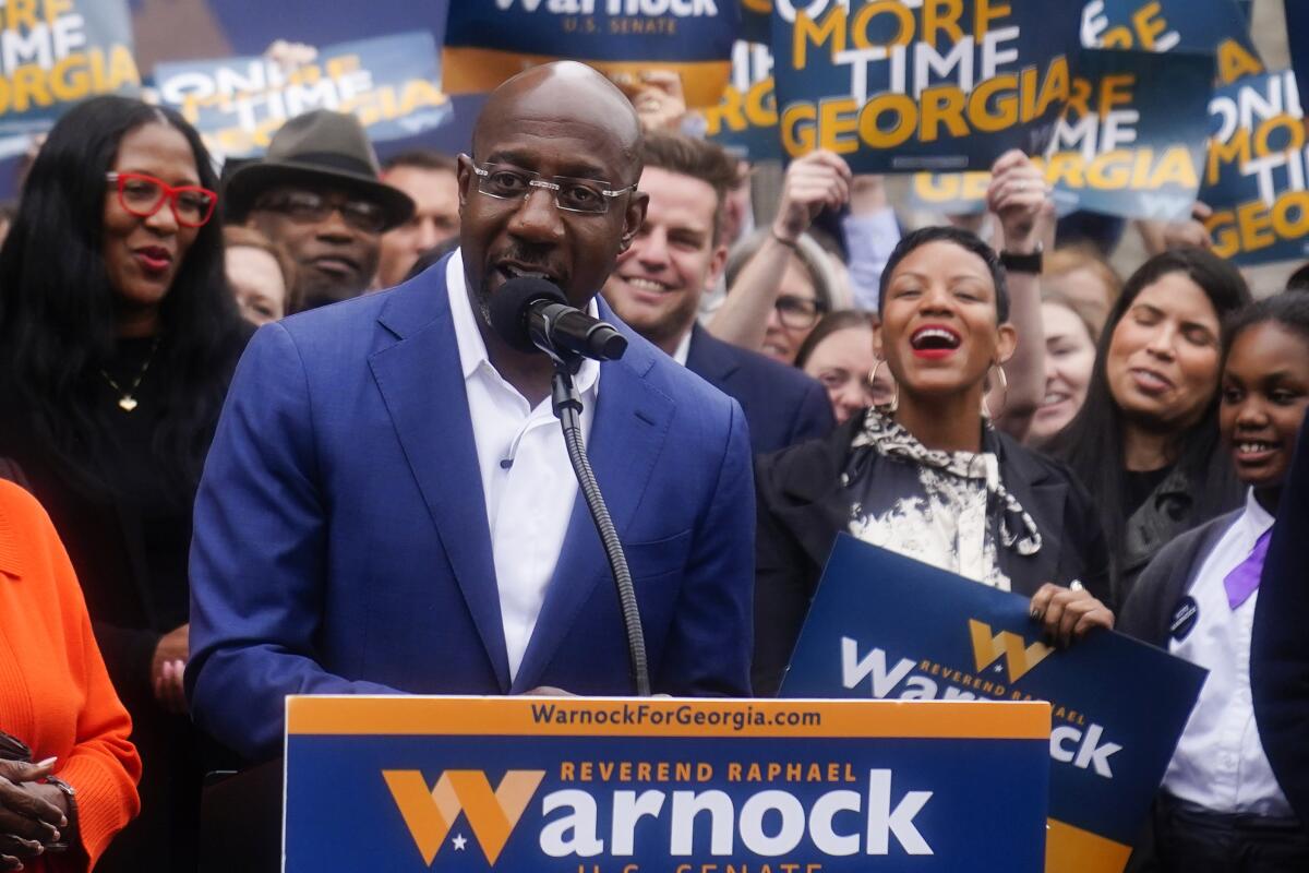 Sen. Raphael Warnock speaks at a lectern surrounded by supporters holding his campaign signs. 