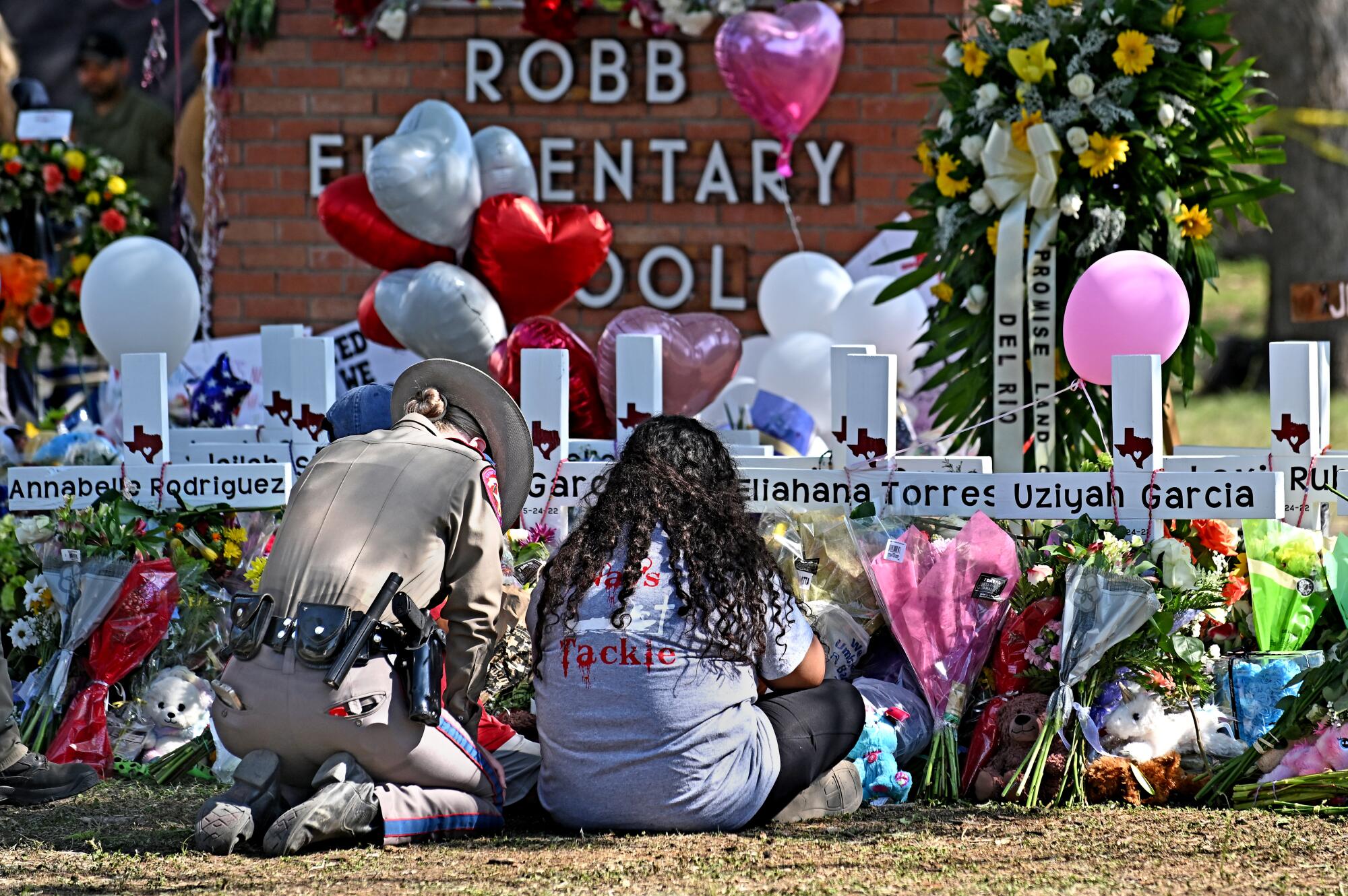 A police officer kneels with a woman at a memorial with flowers and balloons outside a school.