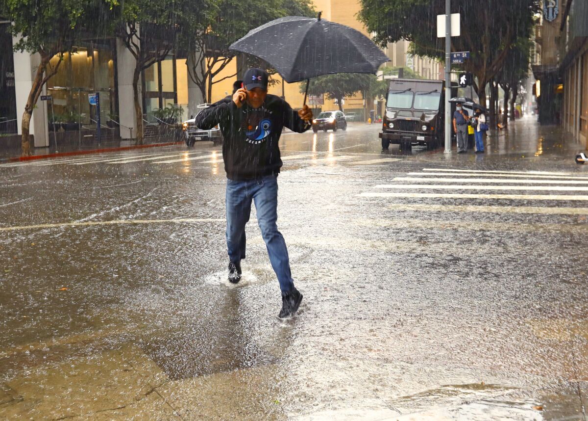 A man with an umbrella attempts to hop over a very large puddle.