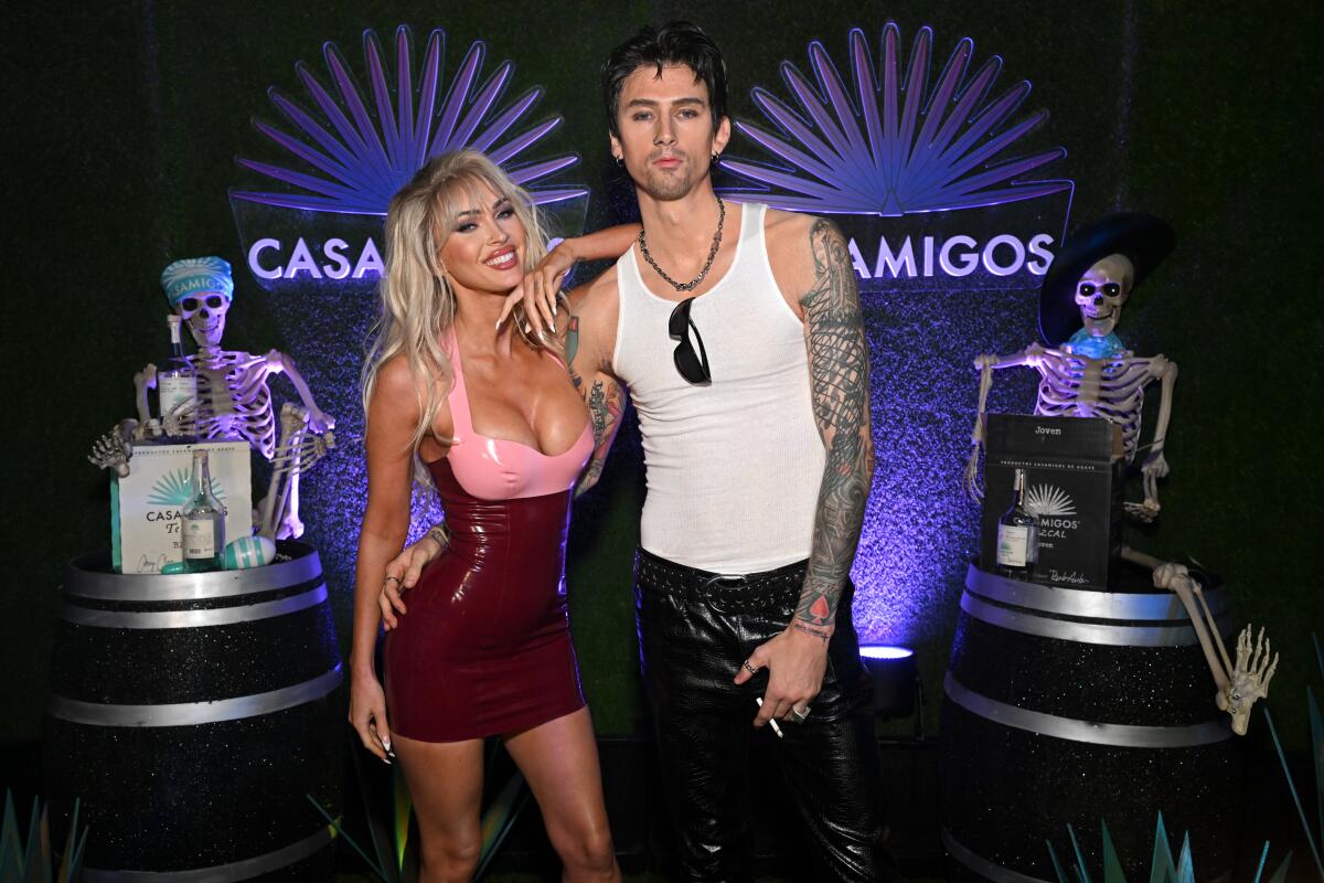 A woman wearing a blond wig and a latex dress posing with a man with lots of tattoos wearing a white tank top