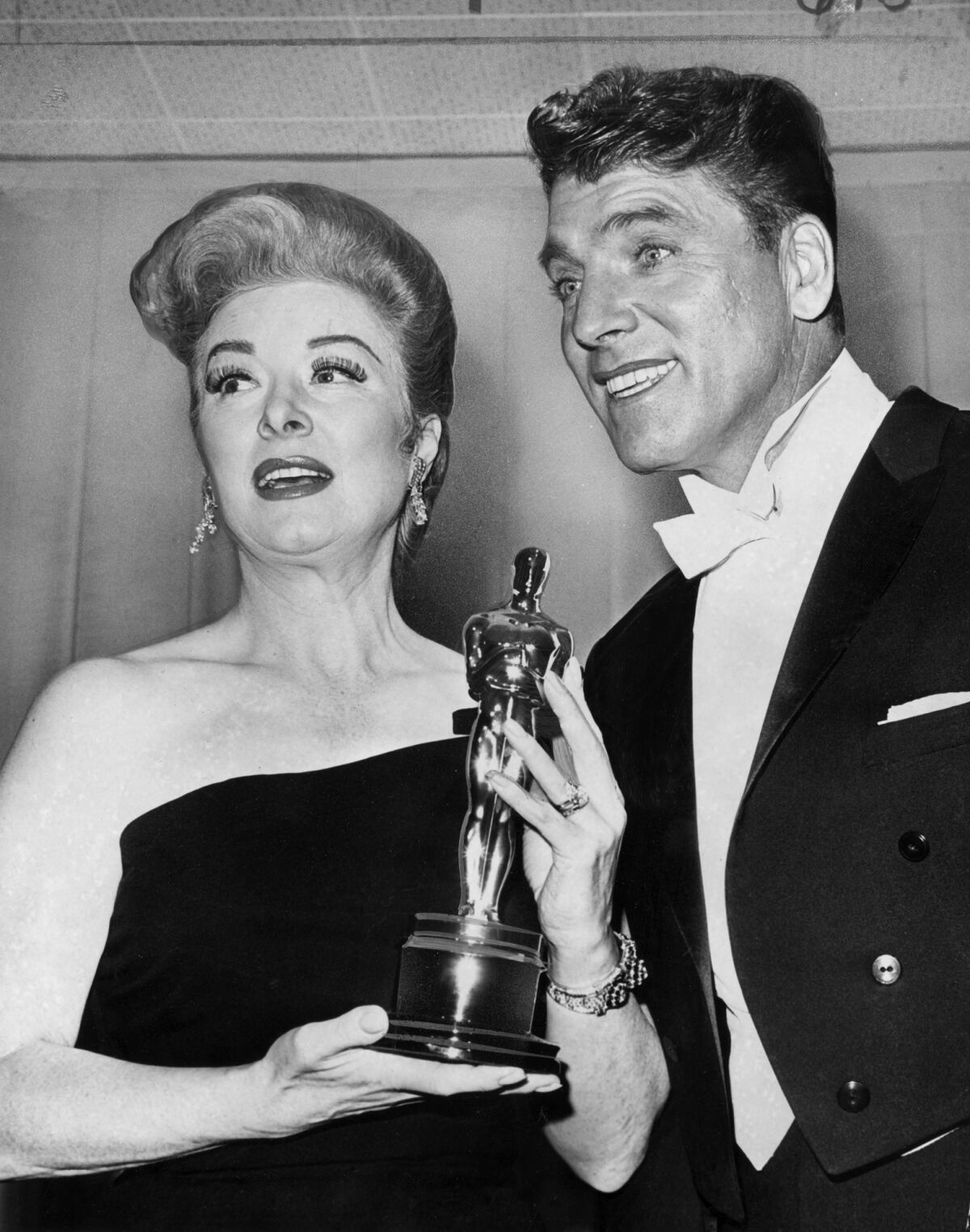 A woman in a strapless gown holds a statuette alongside a smiling man in white tie and tails.