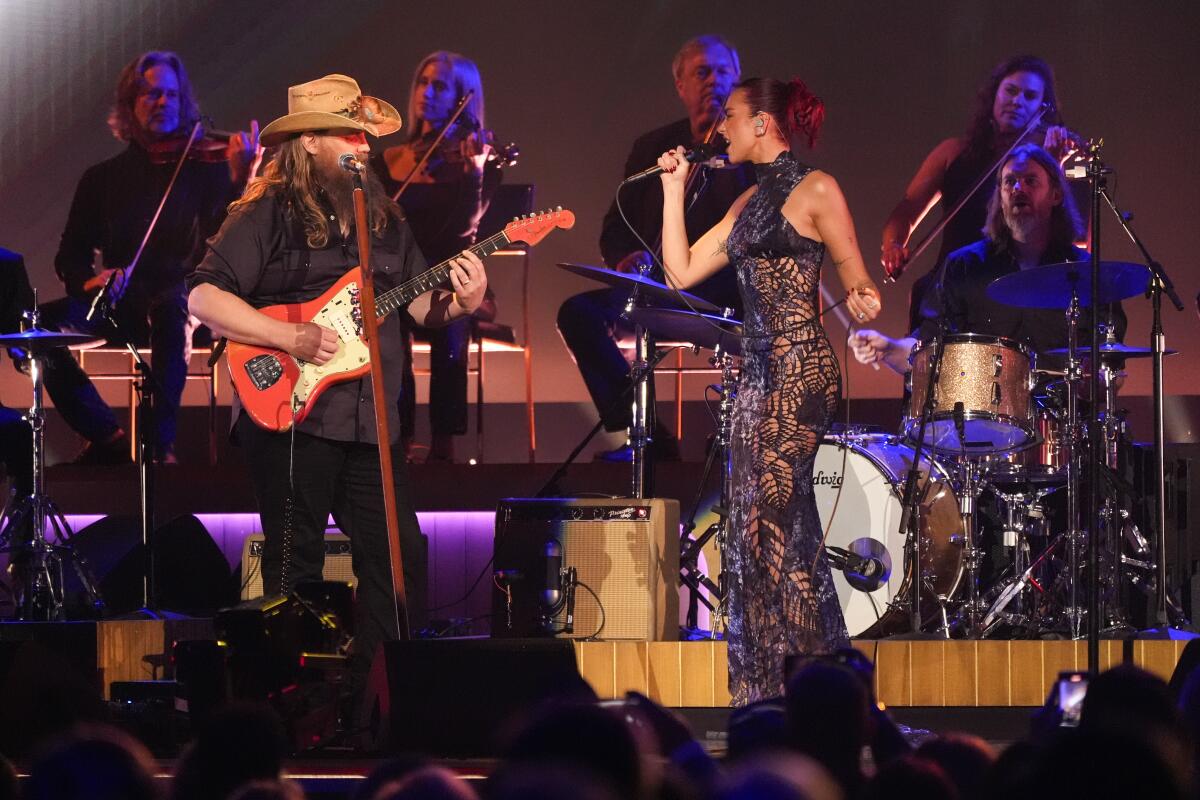 Chris Stapleton, left, wearing a cowboy hat and Dua Lipa in a lacy dress perform onstage