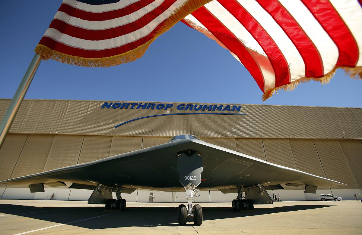 A B-2 stealth bomber is parked in front of a Northrop Grumman facility while the American flag flies overhead.
