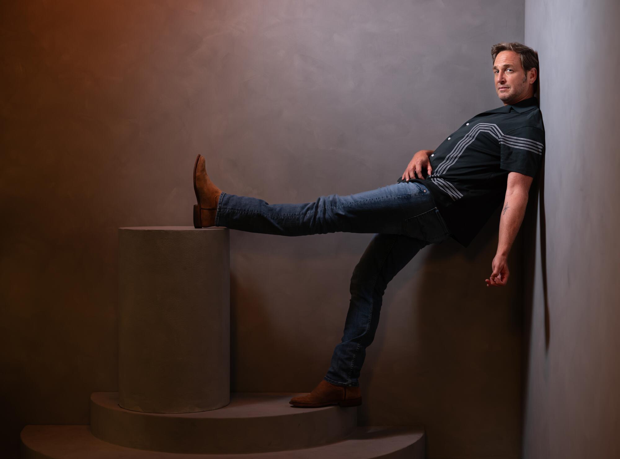 Josh Lucas leans with his back against a wall and his leg stretched out up on a raised block for a portrait.
