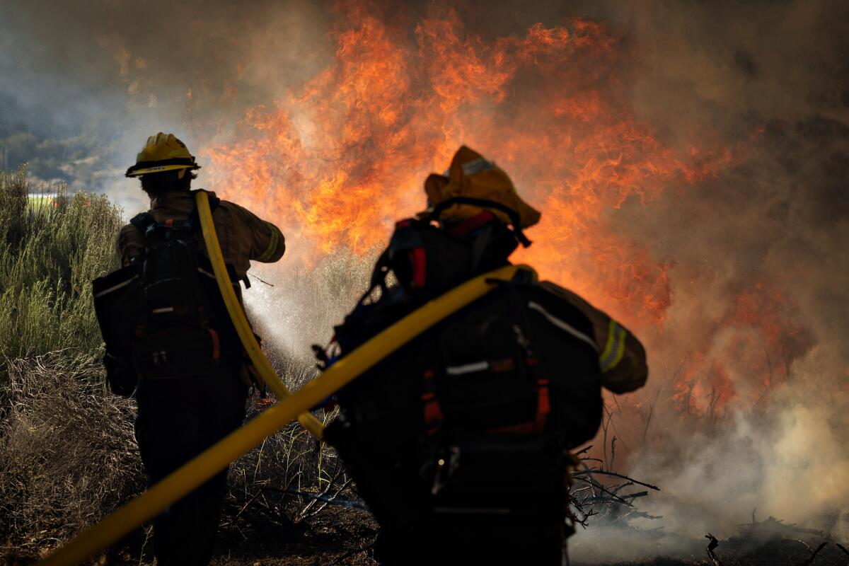 Two firefighters carry a hose toward flames.