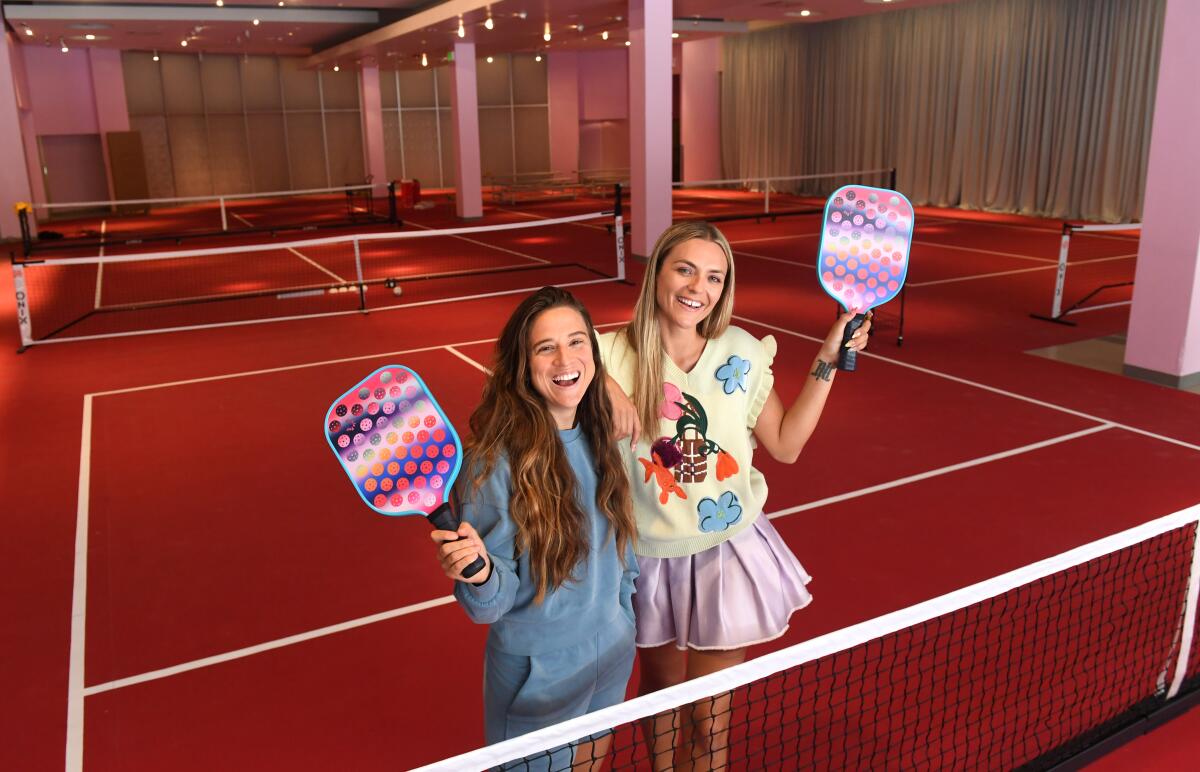 two women smile and hold pickle ball paddles on a red court with white lines and pink walls