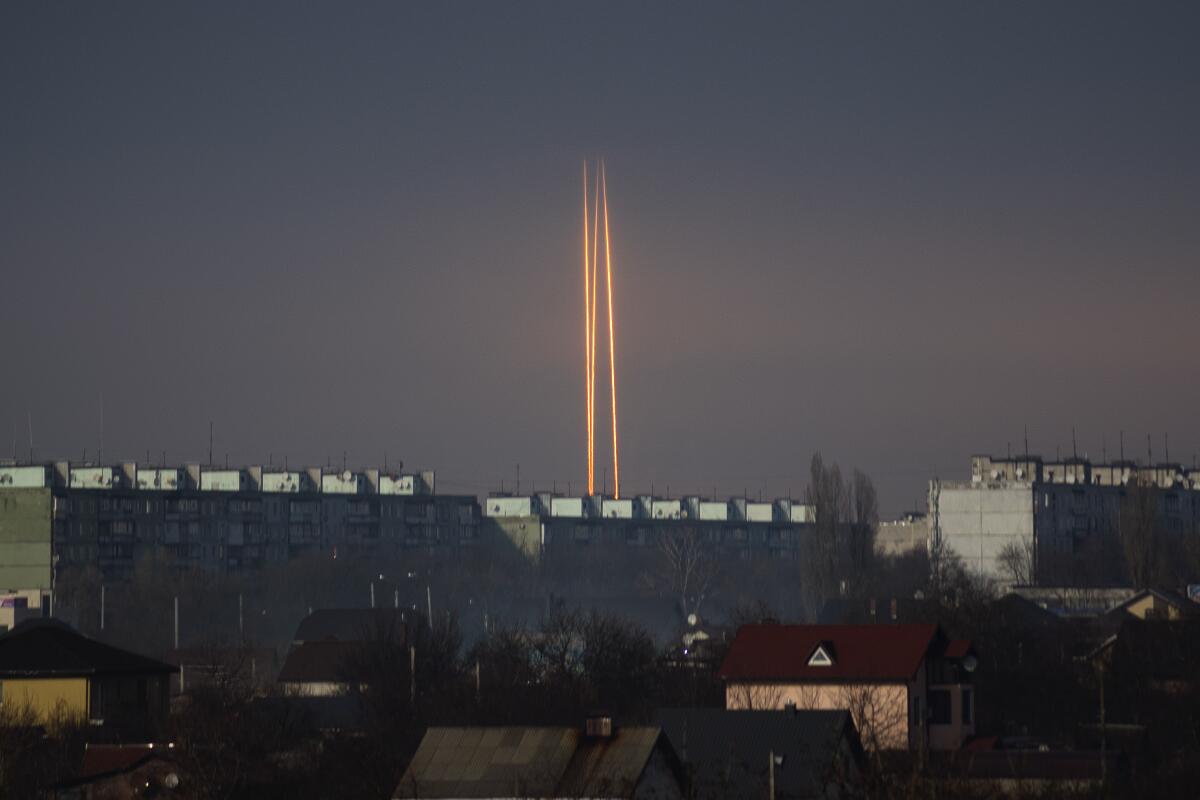Vertical rocket contrails are seen against a dark gray sky, over buildings 