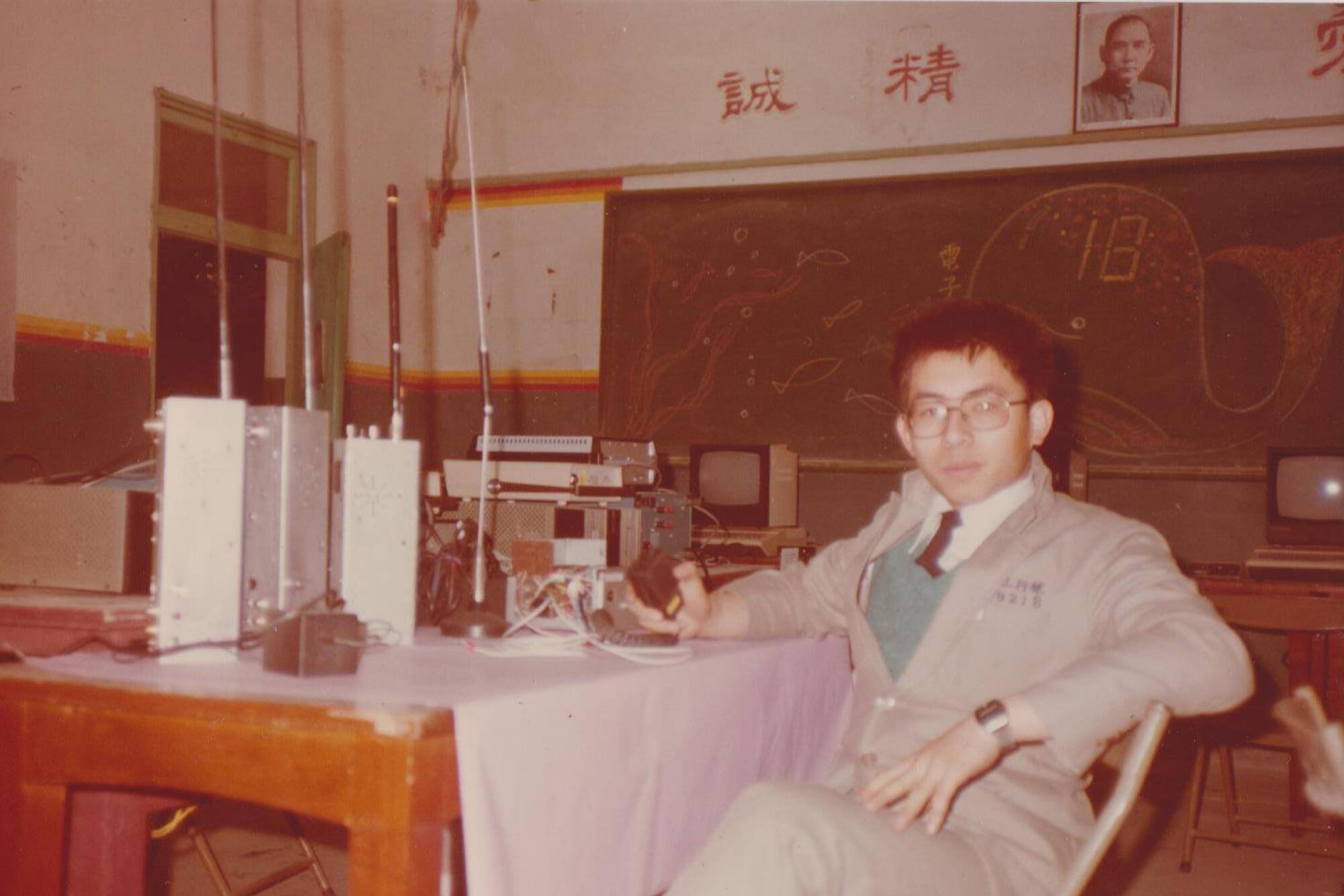 A young man in beige suit and glasses, seated at a table with radio equipment