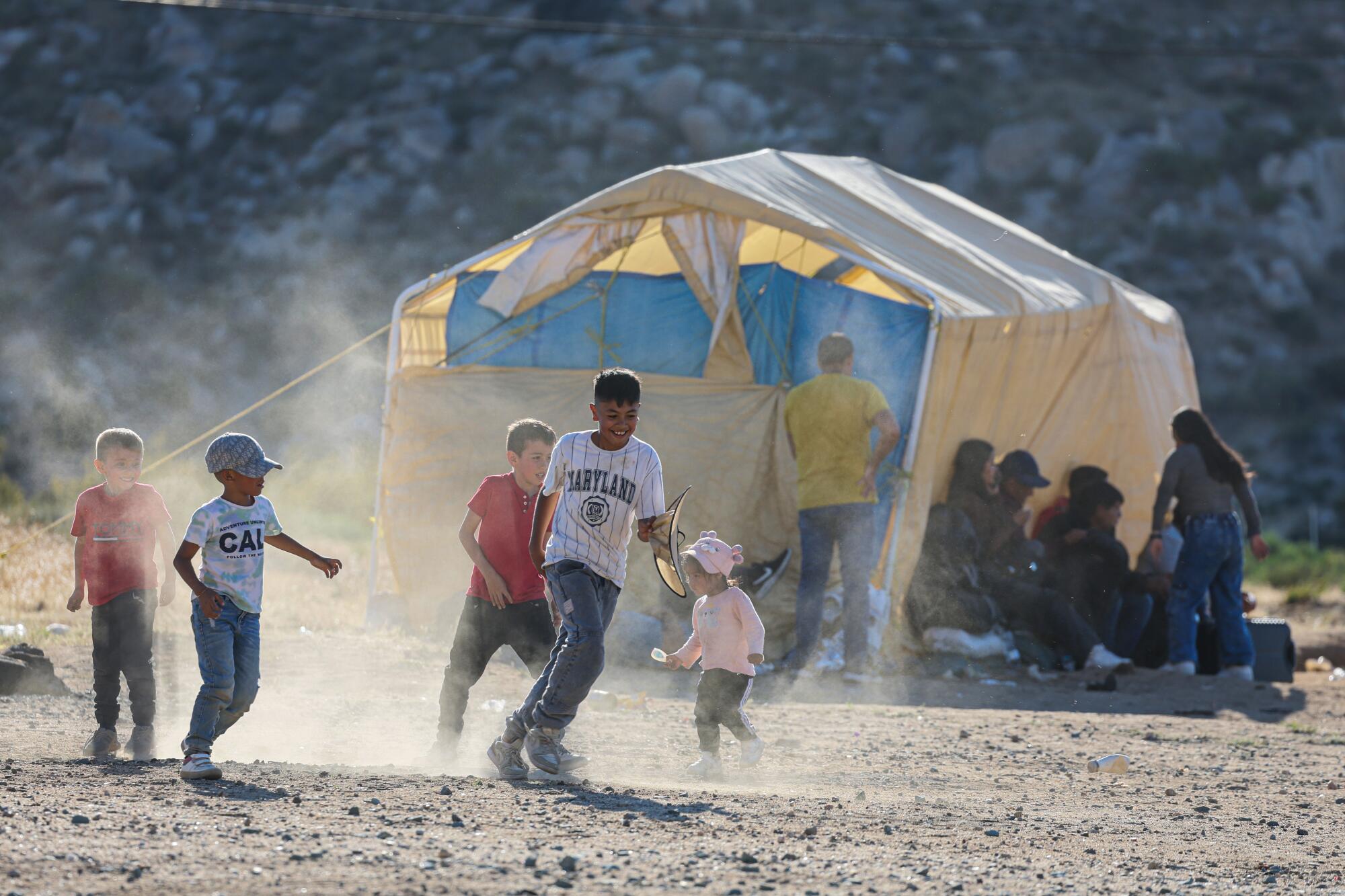 Children play soccer in the dirt with a tent in the background. 