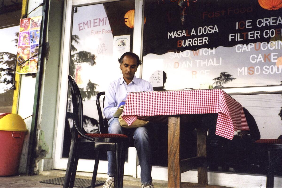 A man sits at a red-check-tablecloth-covered table outside a restaurant.