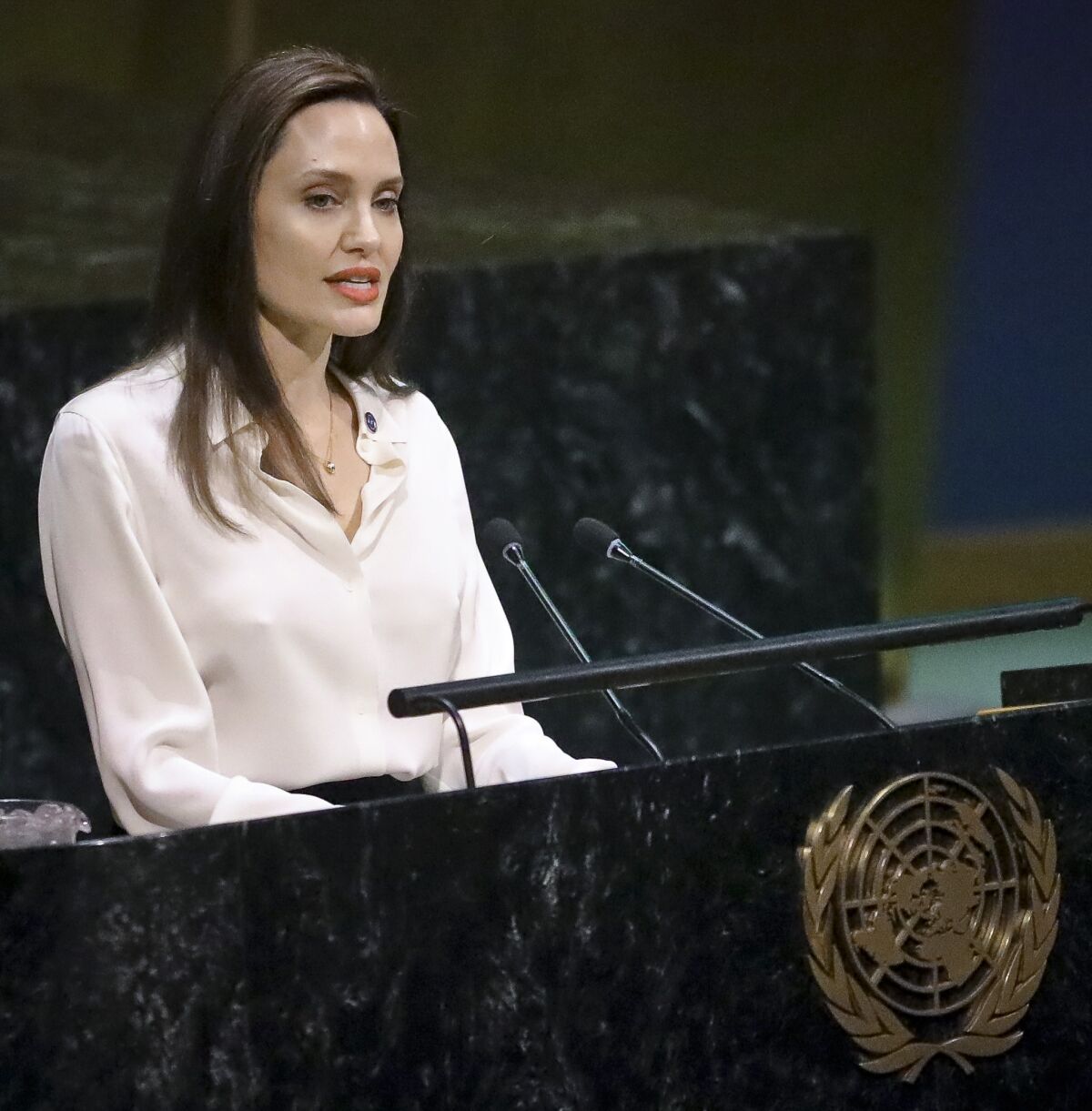 Angelina Jolie in a white blouse speaks into a microphone while behind a United Nations podium