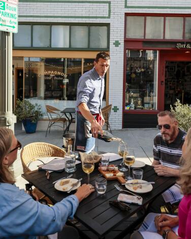 A man pours wine in a glass for a table of patrons nibbling on a cheese plate on an outdoor patio.