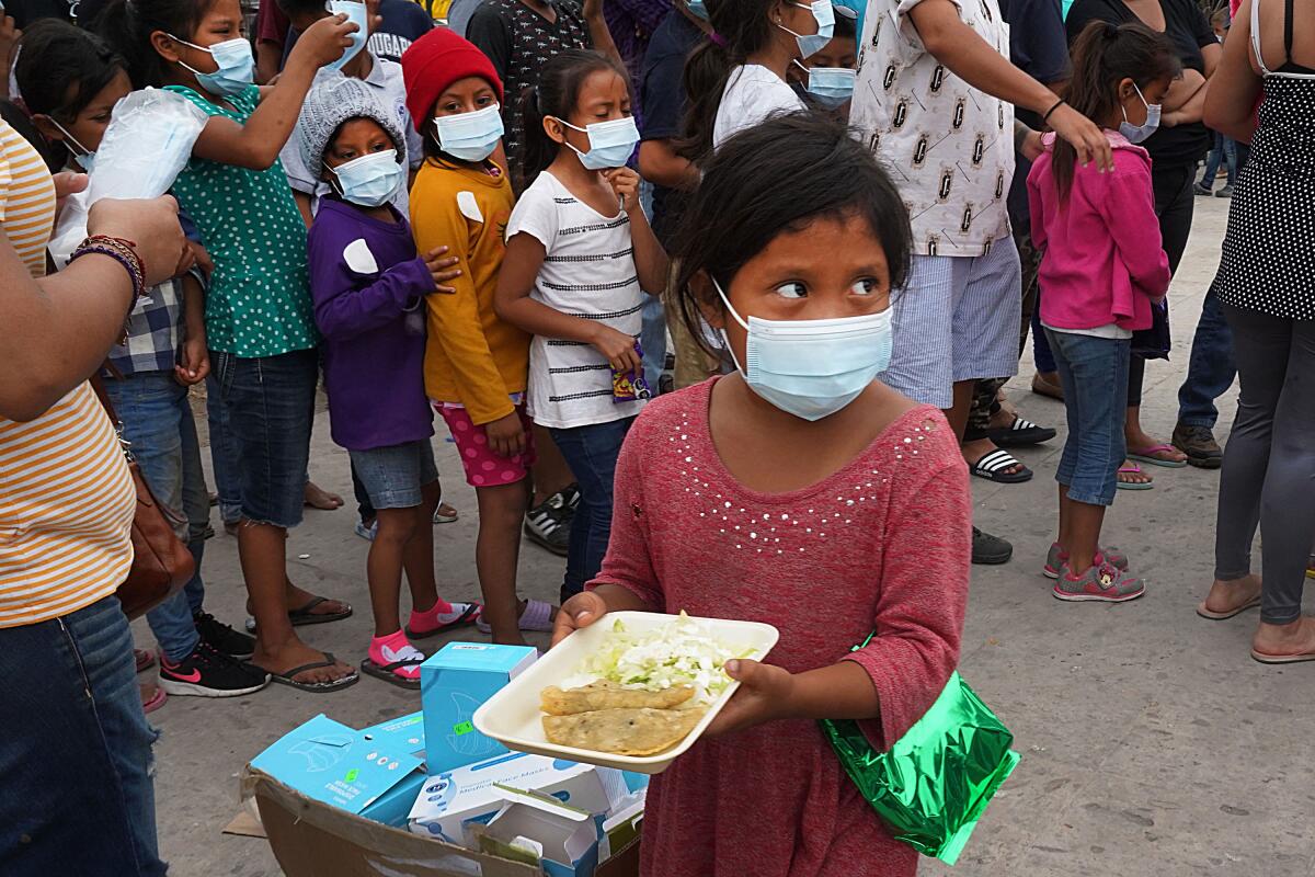 A child holds a plate of food while other children and adults are lined up nearby