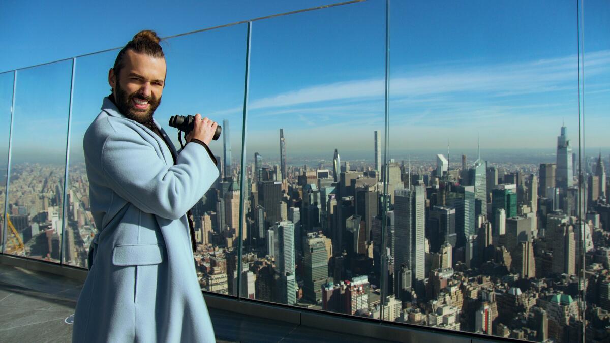 Jonathan Van Ness in a blue coat with binoculars looking out at a city