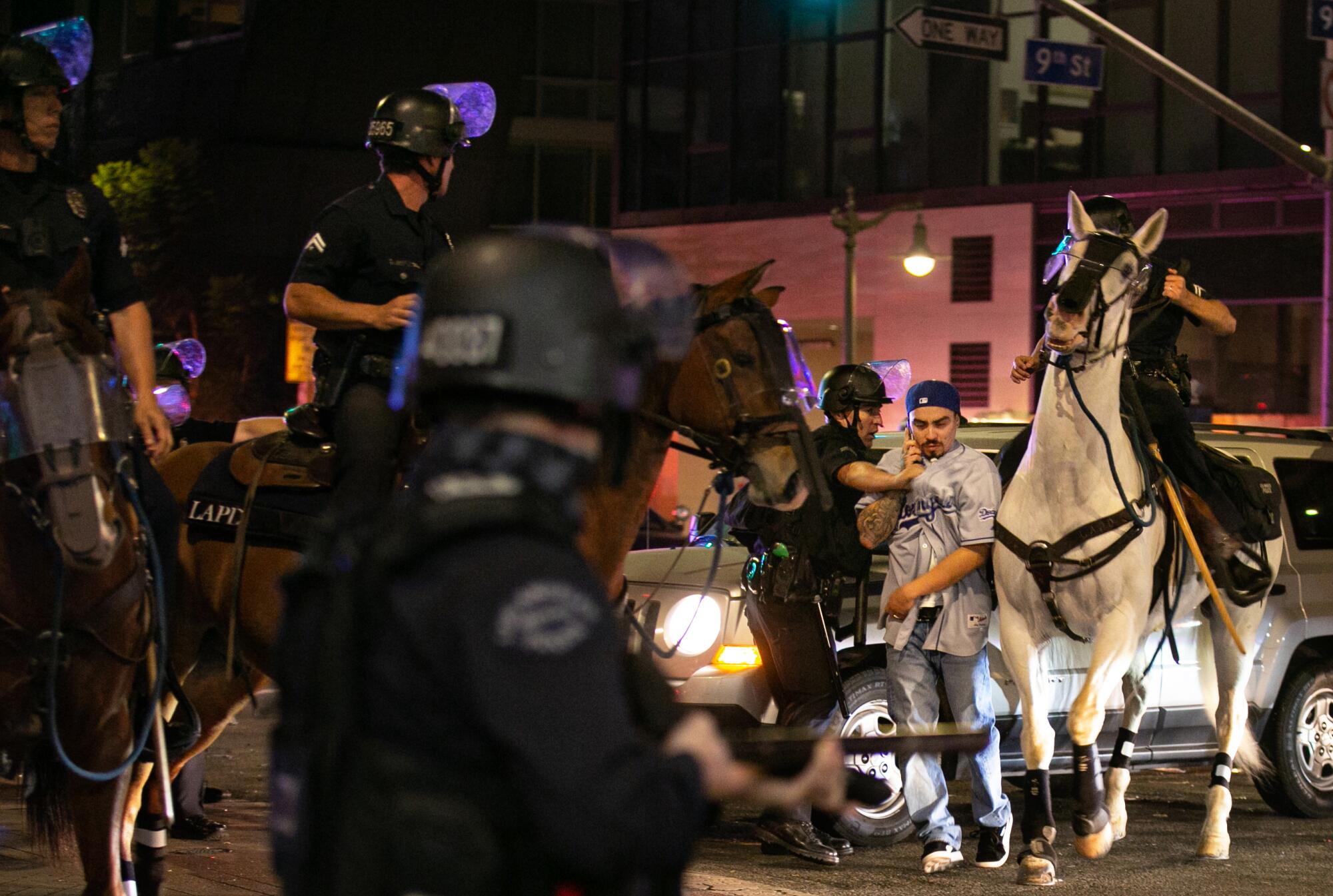 An officer holds a weapon, and another pulls a man in a Dodgers jersey as police on horseback trot past.