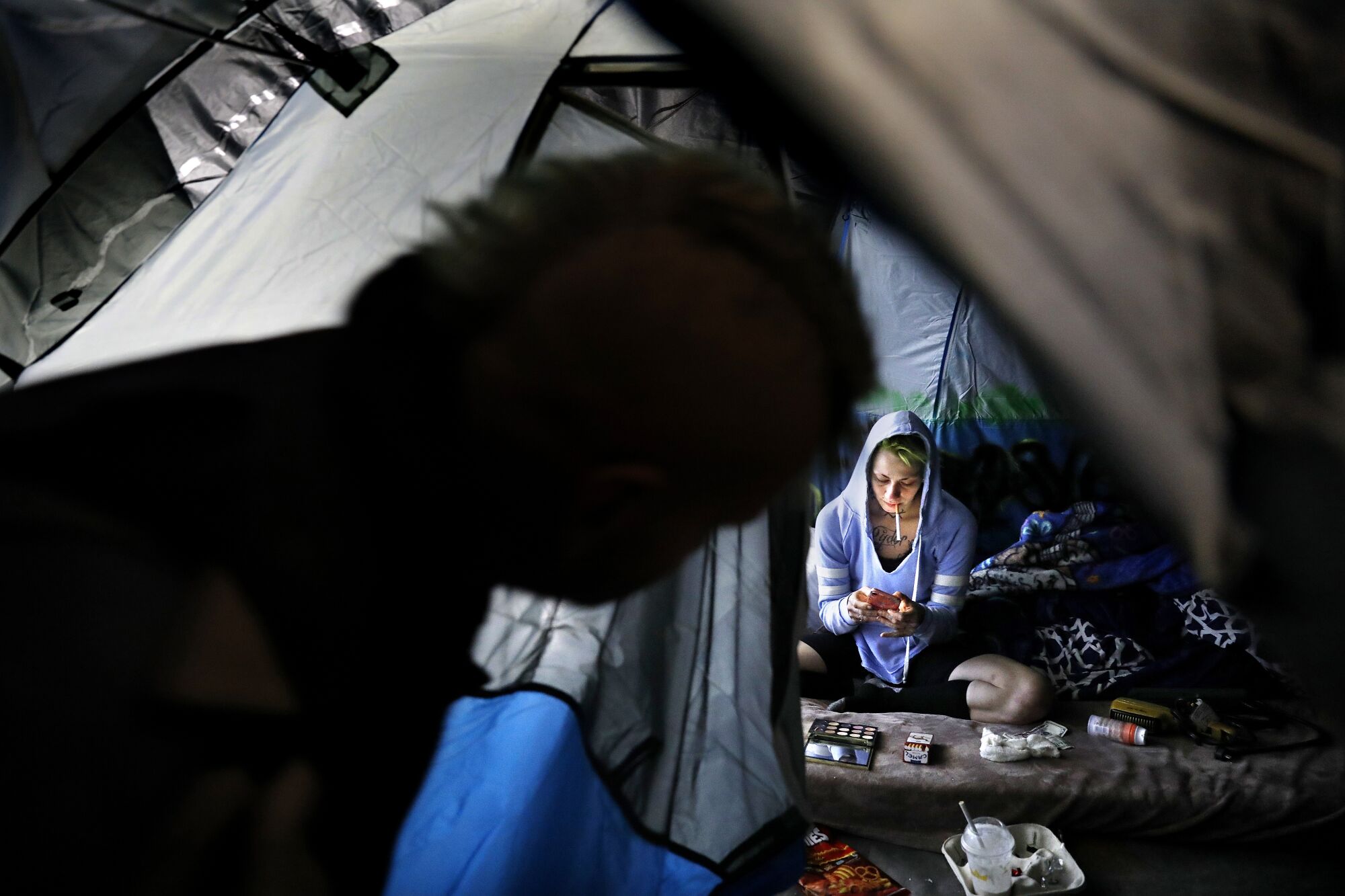 A person in shadow peers into an open tent where a woman in a hoodie smokes and looks at her phone.