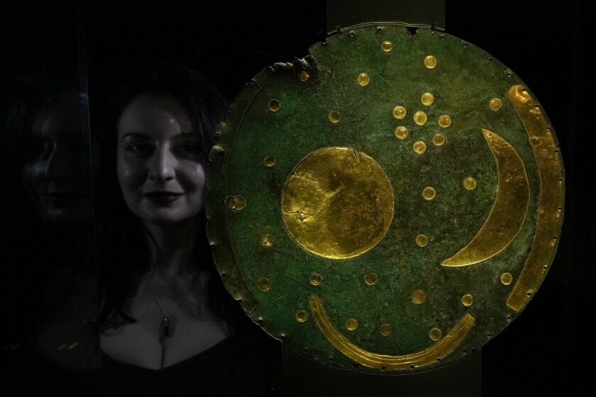 A member of staff poses next to the 'Nebra Sky Disc' which dates from around 1600 BCE, and is the oldest surviving representation of the cosmos, on display at the The World of Stonehenge' exhibition at the British Museum in London, Monday, Feb. 14, 2022. The exhibition which displays objects and artifacts from the era of Stonehenge opens 17, Feb. and runs until 17, July 2022. The Nebra Disc was found in Nebra in Saxony-Anhalt in east Germany, in 1999. (AP Photo/Alastair Grant)