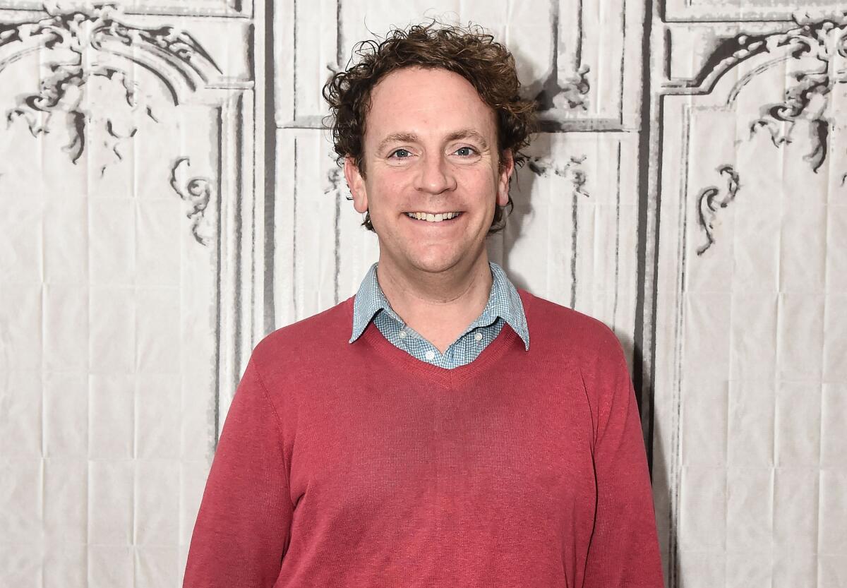 A man with curly hair, wearing a red sweater over a gray collared shirt, stands smiling in front of a white wall.