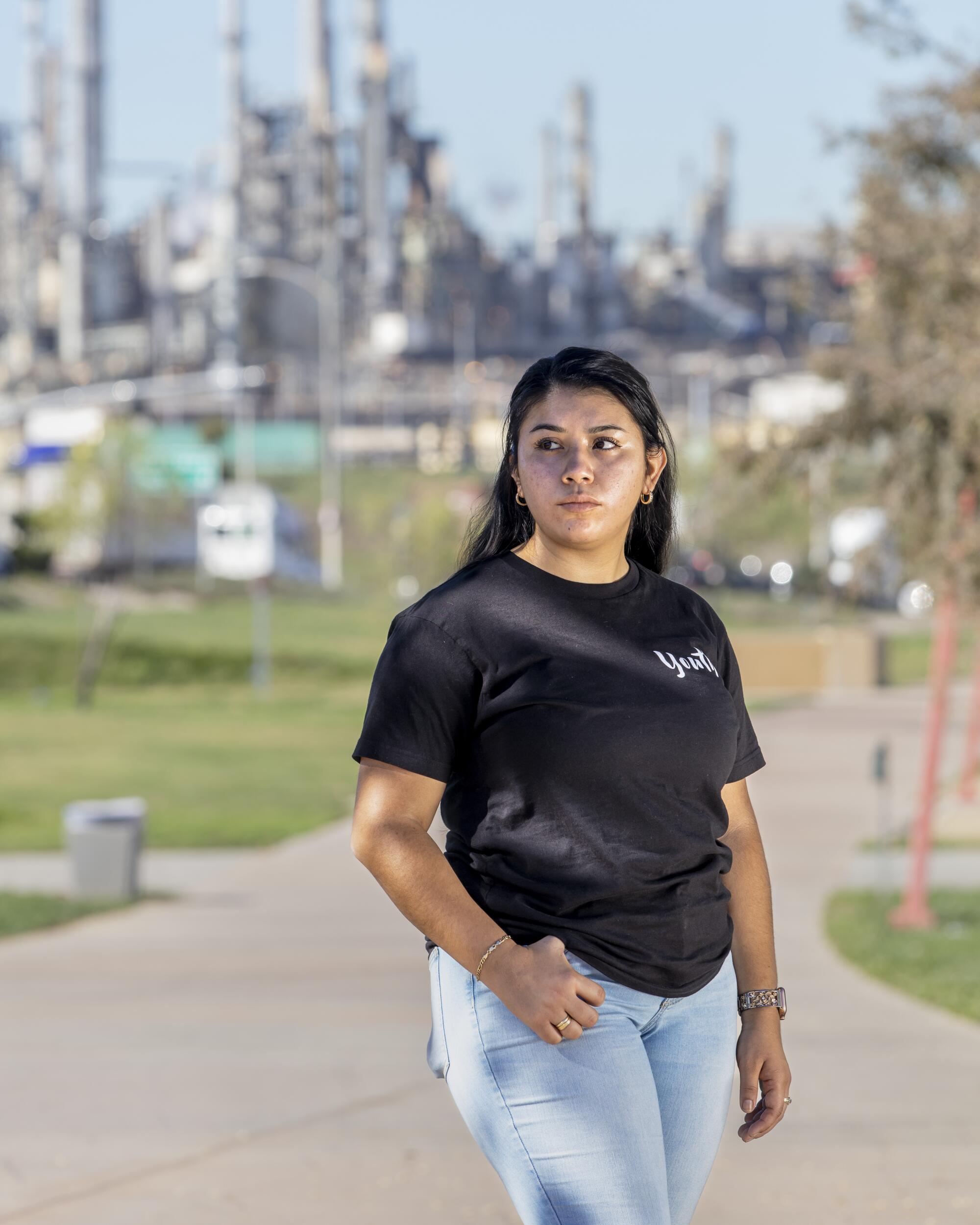 A young woman stands with an oil refinery in the background