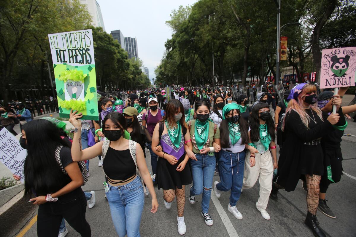 Women and girls march on a street, some wearing green scarves and holding up signs 