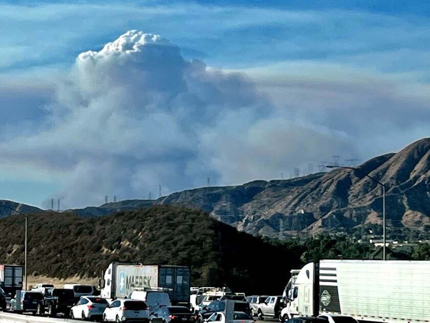 A huge cloud of smoke rises above mountain peaks as vehicles sit in traffic on a highway in the foreground
