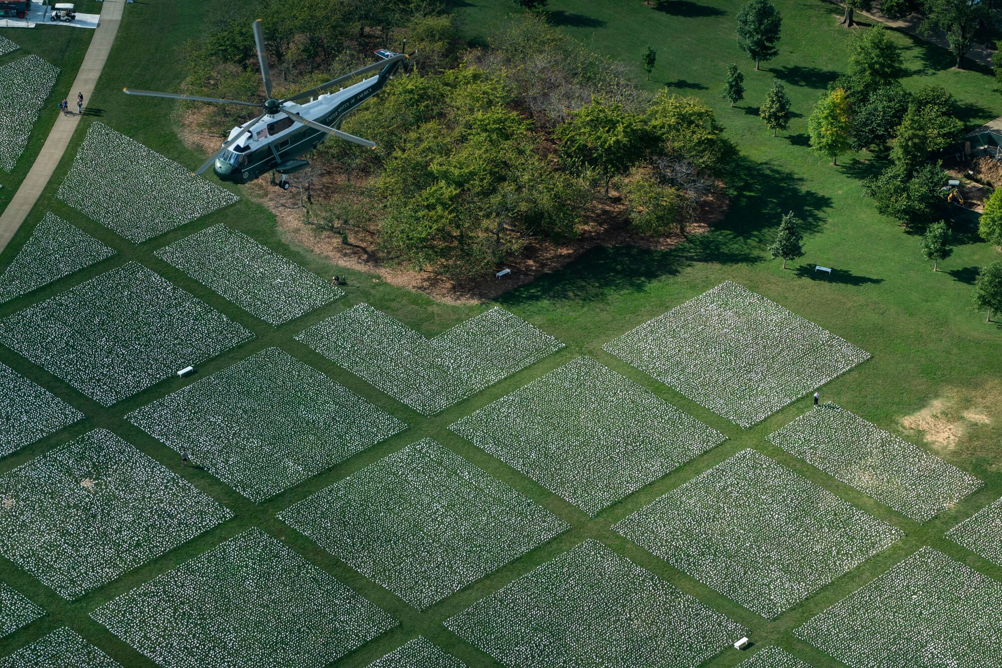 Helicopter flies over the National Mall, with grids of small white flags planted in the grass