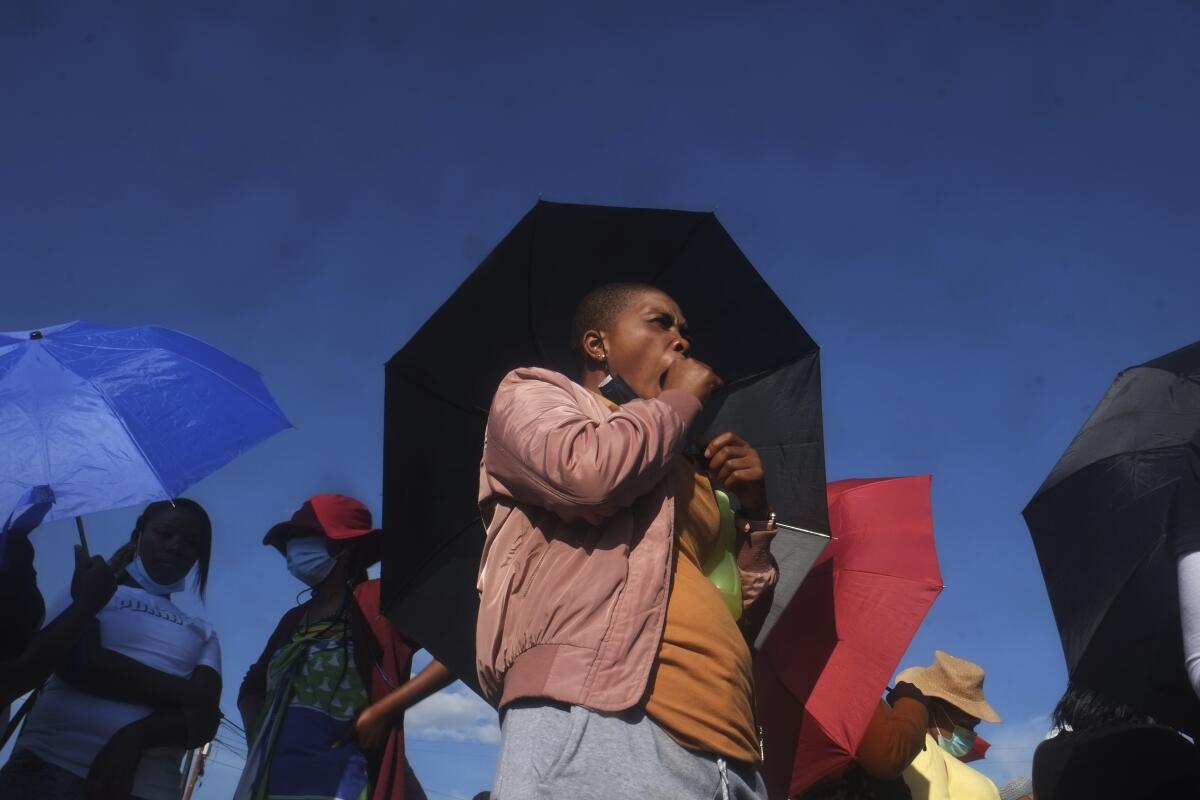 A person with short hair and pink jacket, holding a black umbrella, yawns while standing among other people 