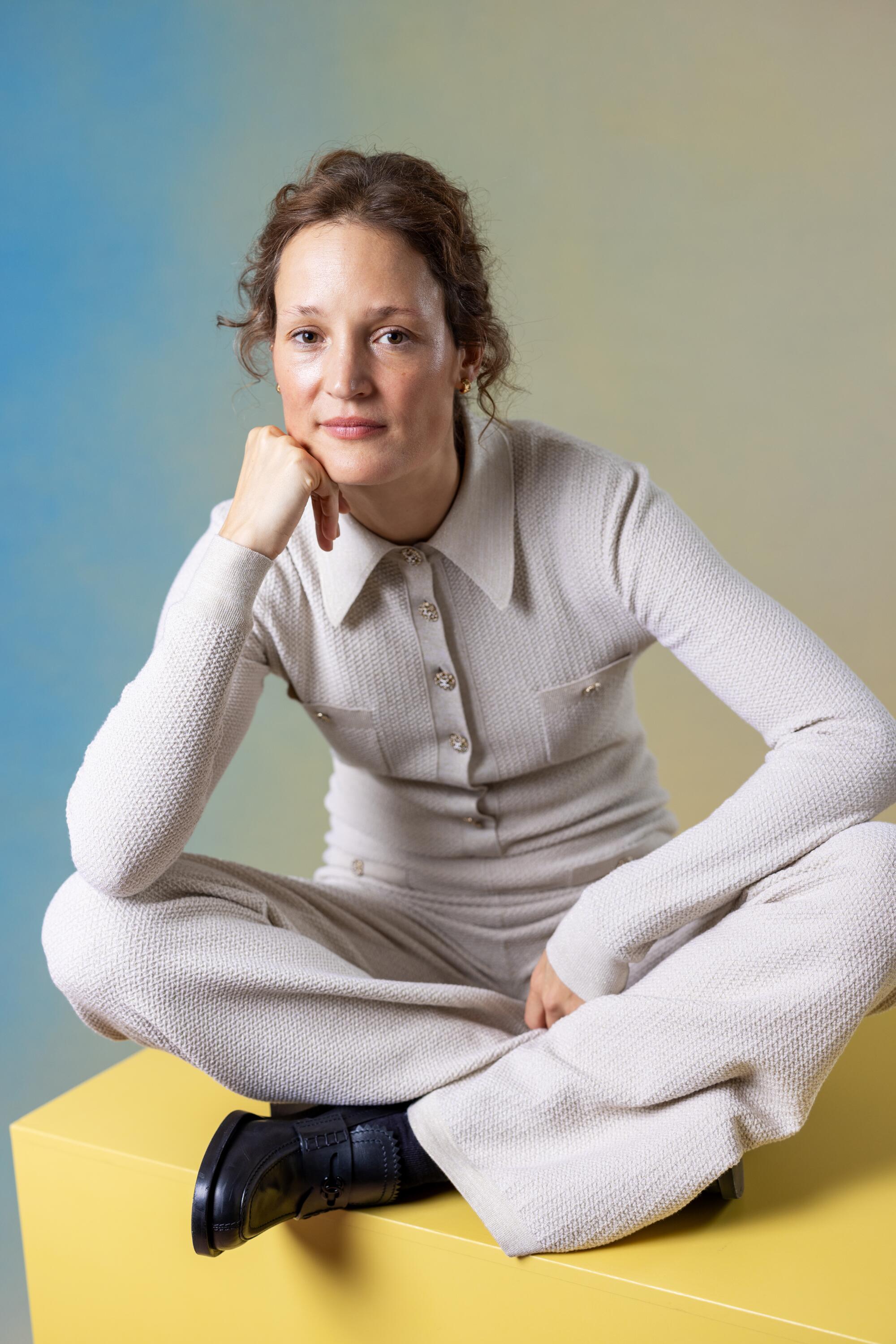 Vicky Krieps sits cross-legged in a white outfit on a cube.
