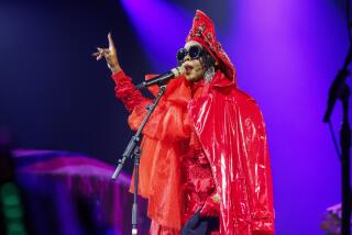 A woman in a red latex coat and large sunglasses on a stage singing into a microphone with her right hand up