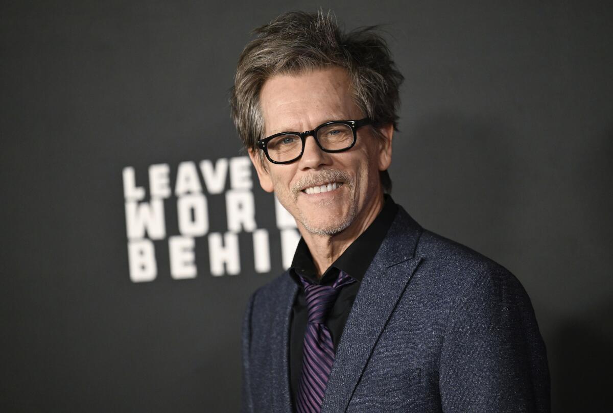 Kevin Bacon smiles in in a dark gray suit and black glasses in front of a background with the words "Leave the World Behind"