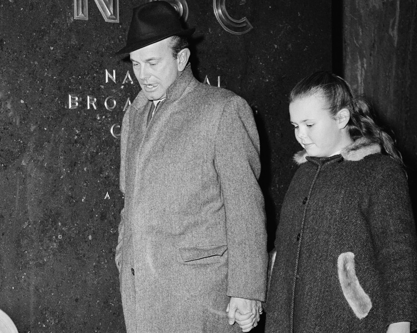 On a city street, a man in a fedora and overcoat holds the hand of a girl in a winter coat with fur collar. 