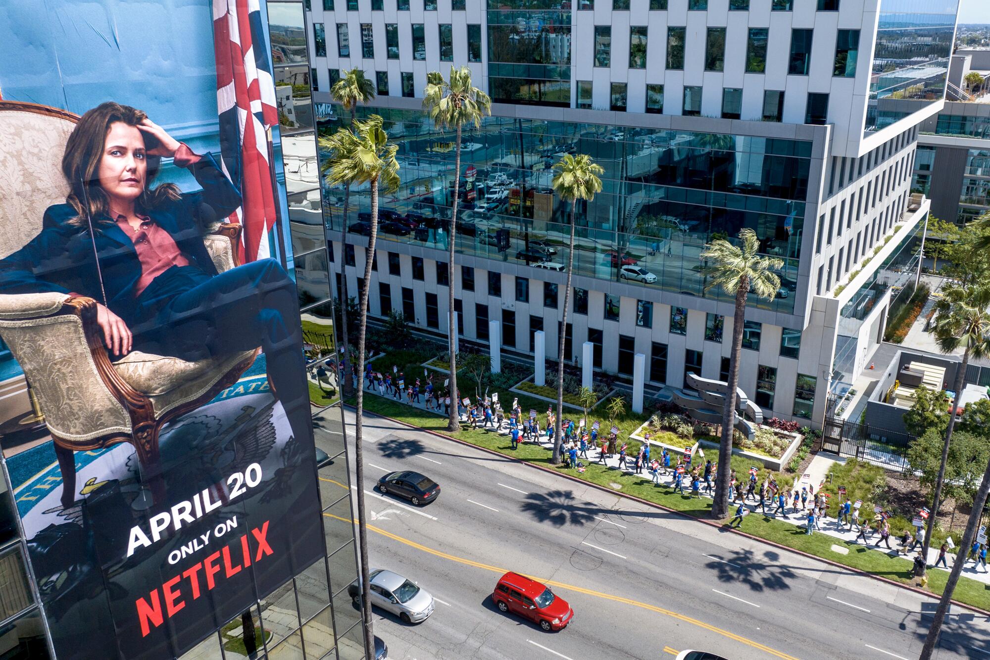 A billboard for a Netflix show hangs on a building across the street where protesters walk a picket line along a building