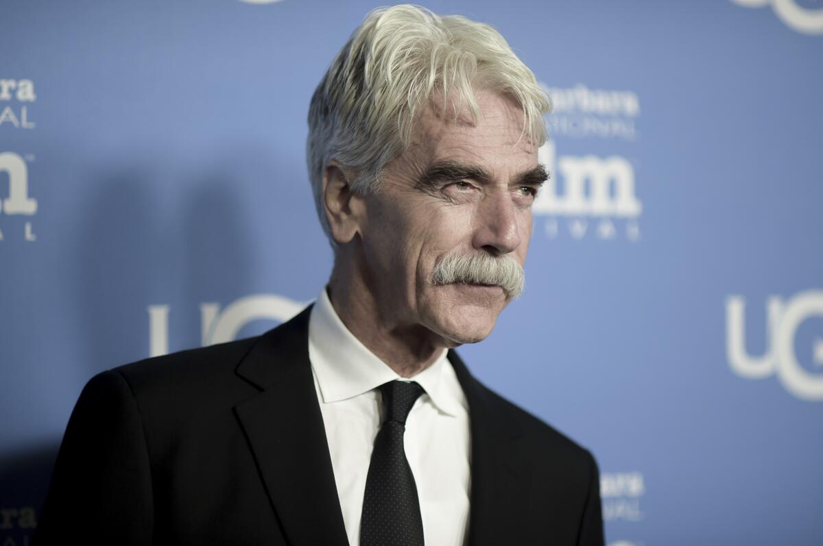 Sam Elliott poses for cameras in a black suit and tie and a white shirt