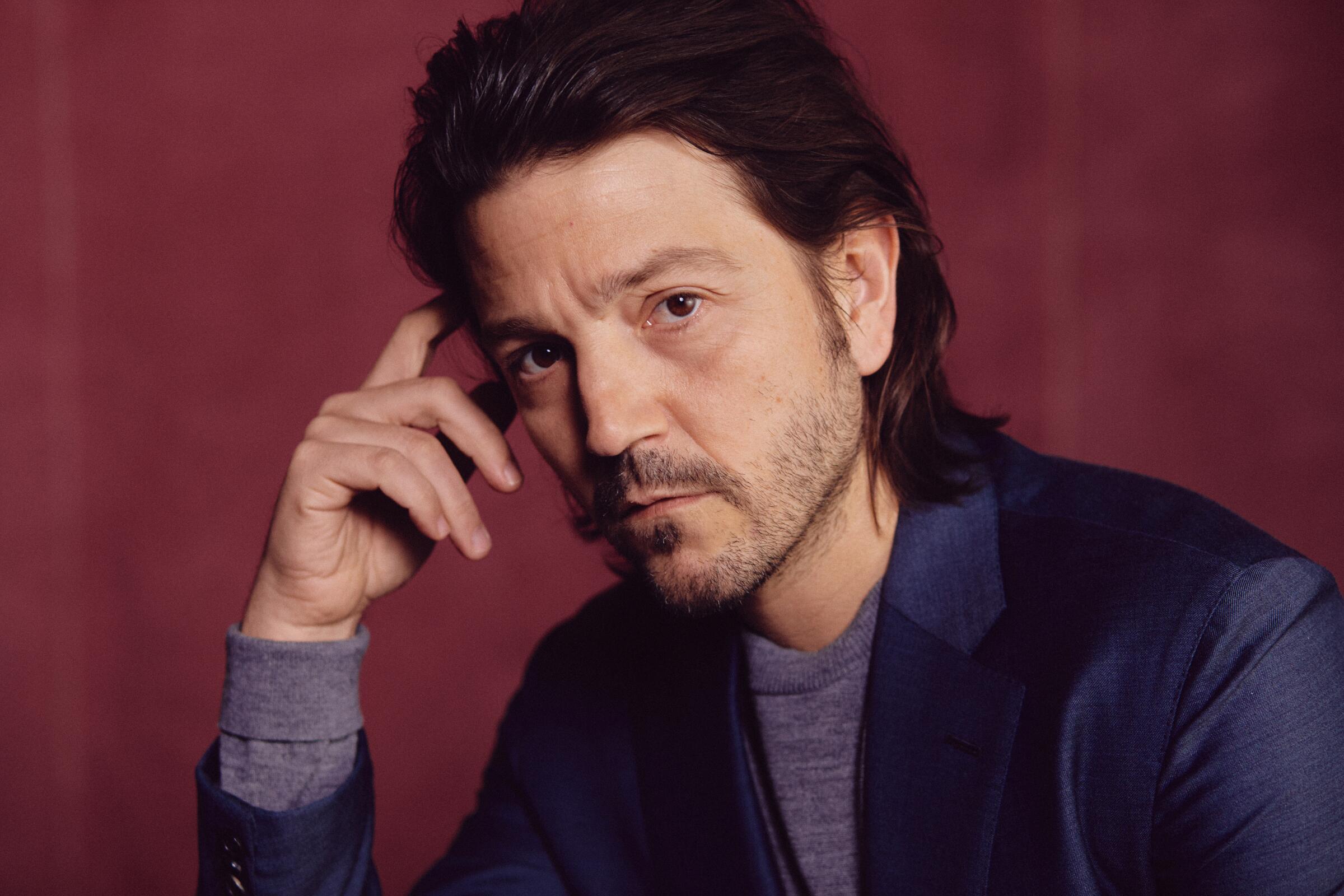 A serious-looking man with scruffy facial hair with his hand held up to his temple