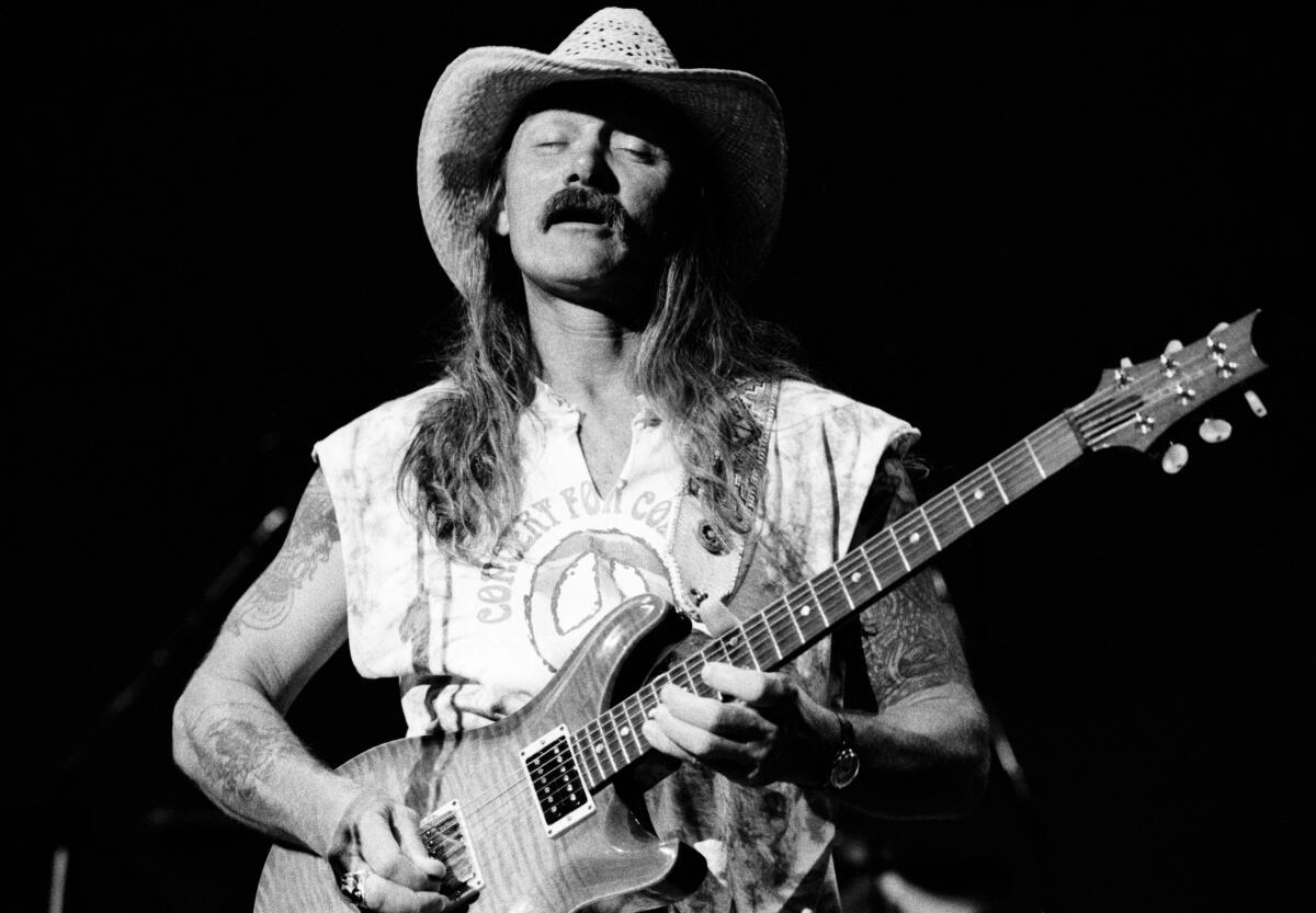 A man with long hair, a mustache, a Grateful Dead shirt and a cowboy hat playing guitar onstage in a black-and-white image