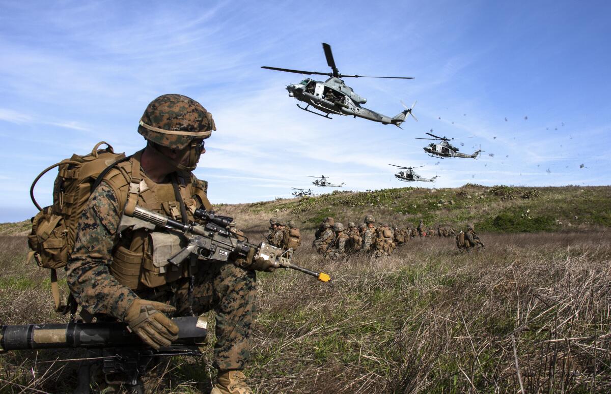 Helicopters fly over Marines in camouflage on the ground.
