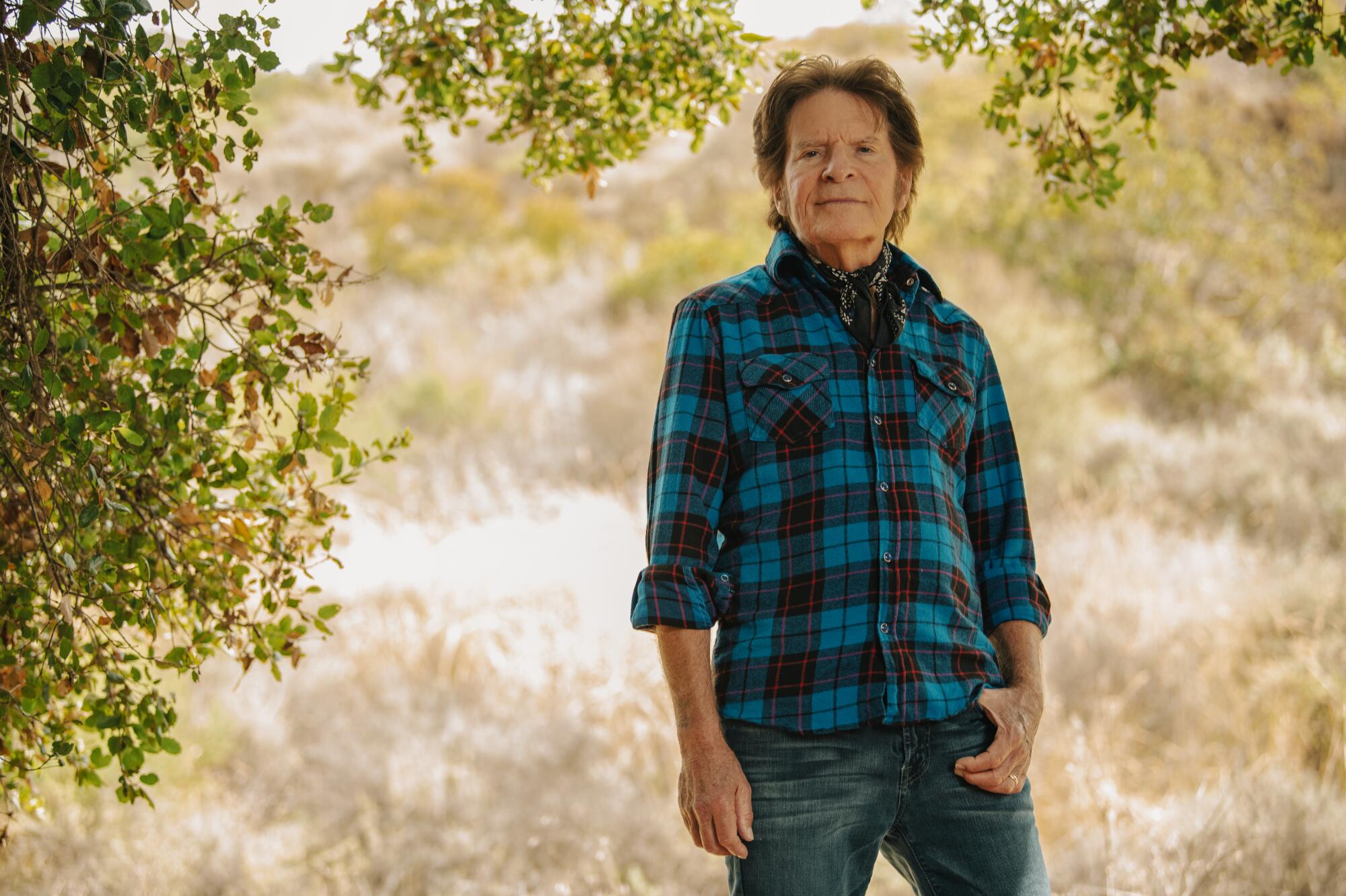 A rock musician in a blue flannel shirt stands in a field