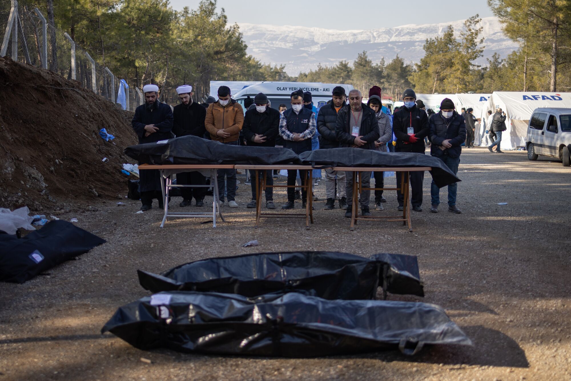 A short row of men stands in prayer over a few black plastic body bags. Snowcapped mountains are on the horizon.