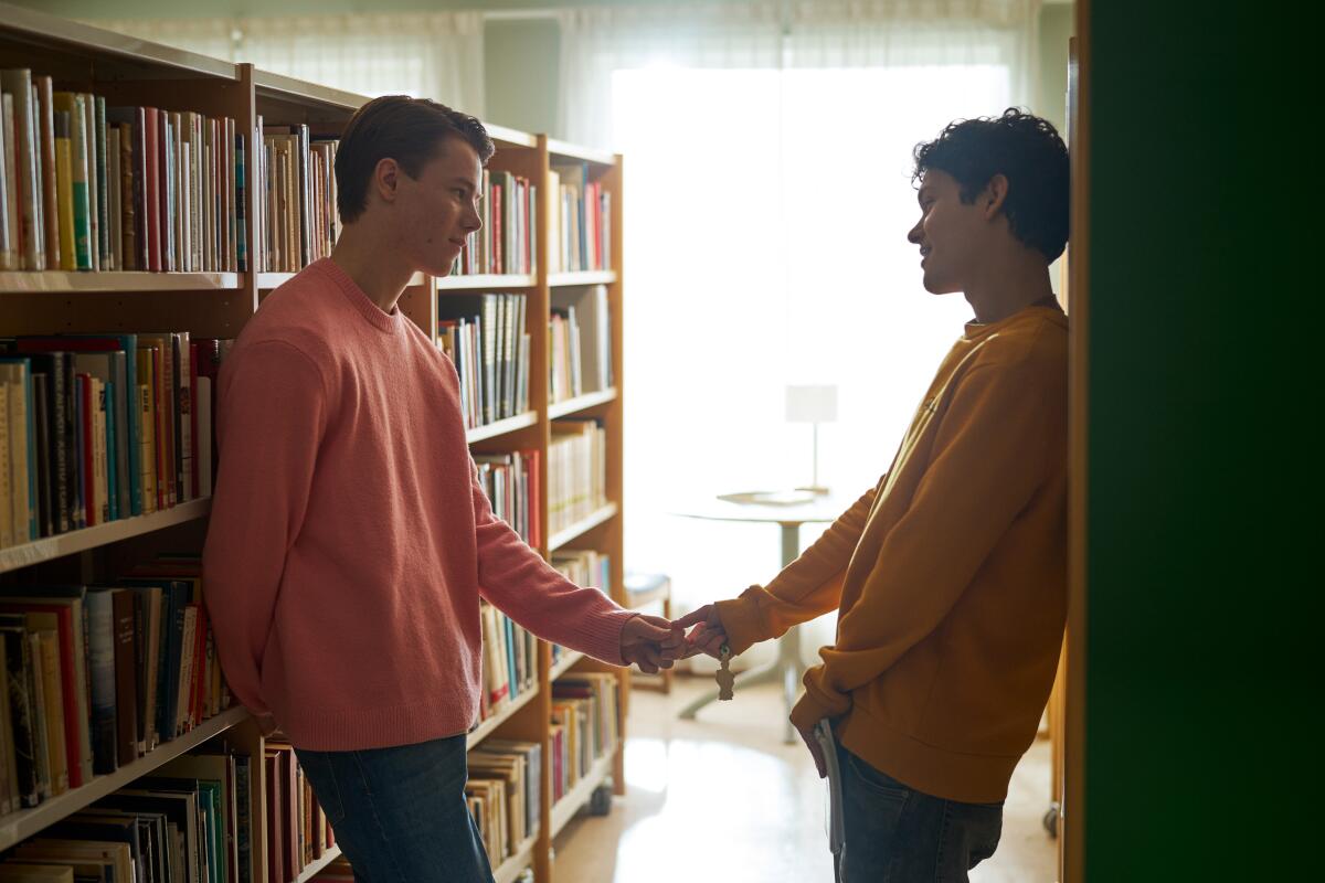 Two boys hold hands as they lean against library bookshelves.