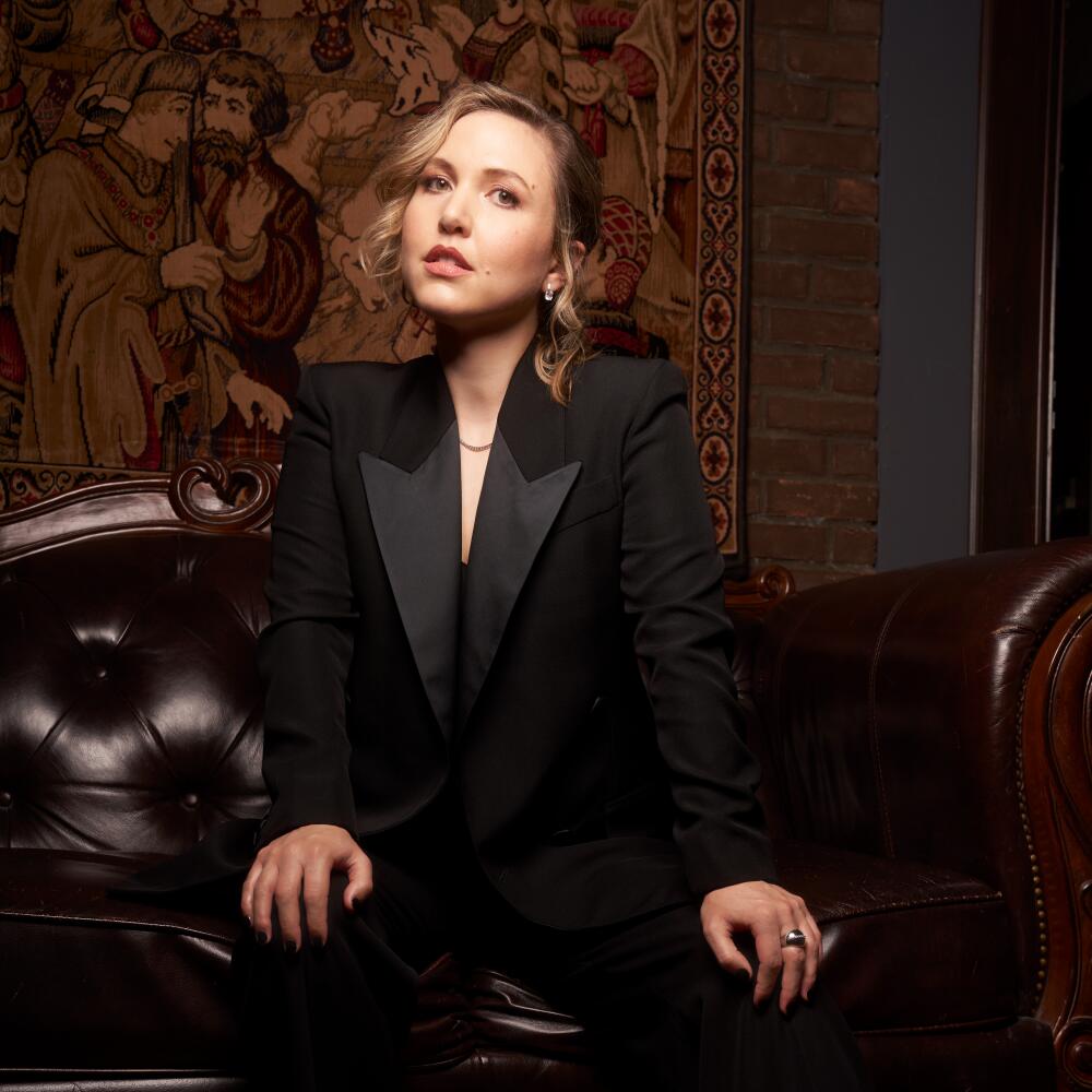 Comedian and late-night host Taylor Tomlinson sits on a leather armchair wearing a tuxedo