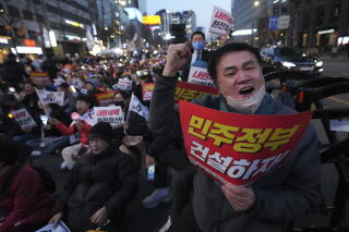 A man raises a fist as he speaks while holding a red sign, joined by a large crowd also holding signs in the street
