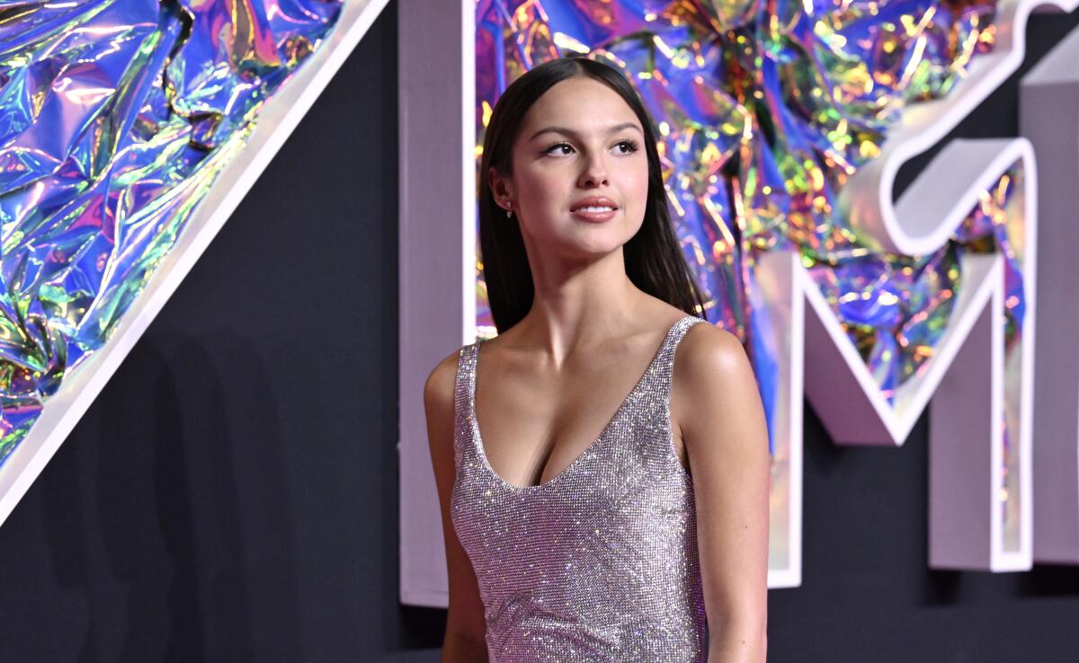 Olivia Rodrigo in a shimmering sleeveless gown posing by looking over her shoulder in front of a backdrop