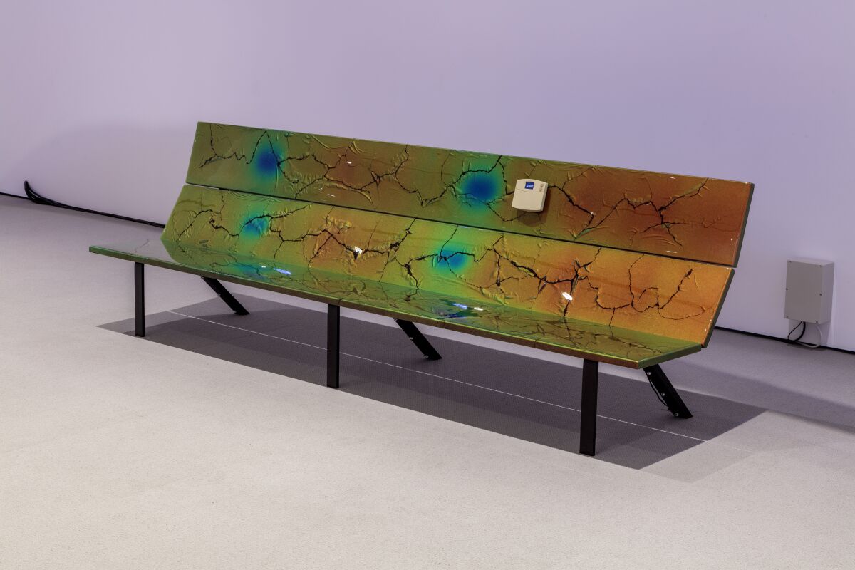 A greenish park bench in a gallery, with painted-on cracks and a sensor