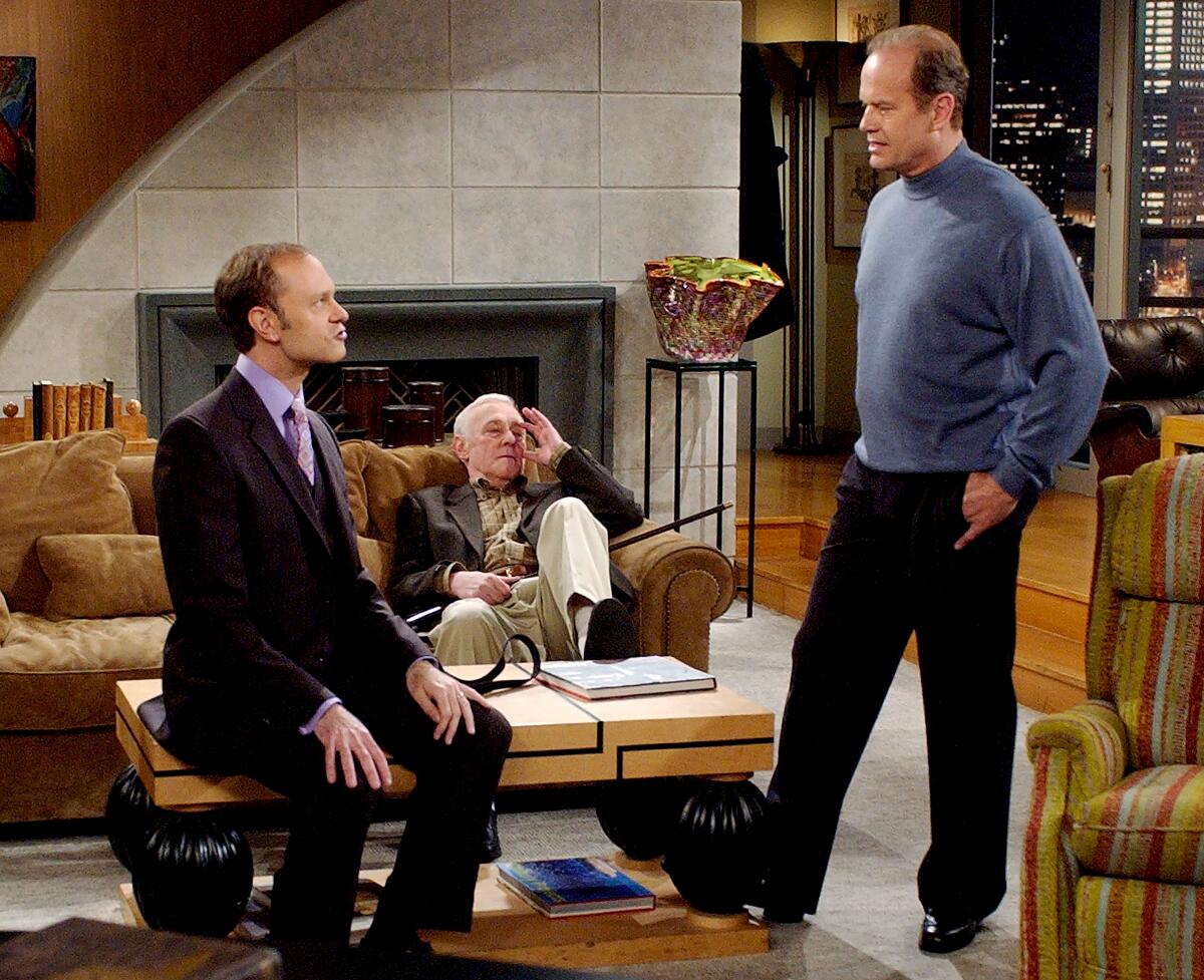 Niles, in a suit, sits on a coffee table looking up at Frasier. Martin sits on a couch behind them and looks at them.