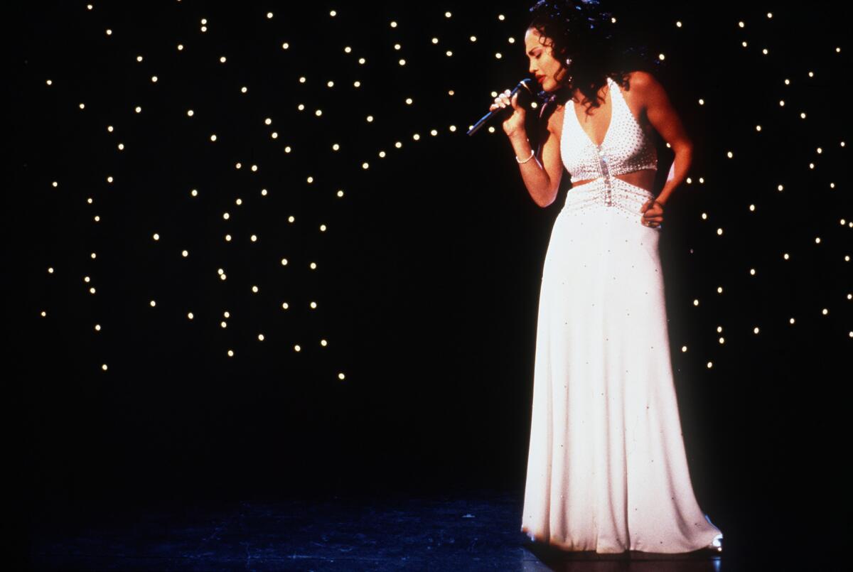 Jennifer Lopez, as Selena, sings on a dark stage in a long white dress.
