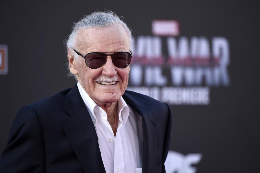 Stan Lee wearing a blue suit and white shirt with no tie and sunglasses smiles at red carpet event