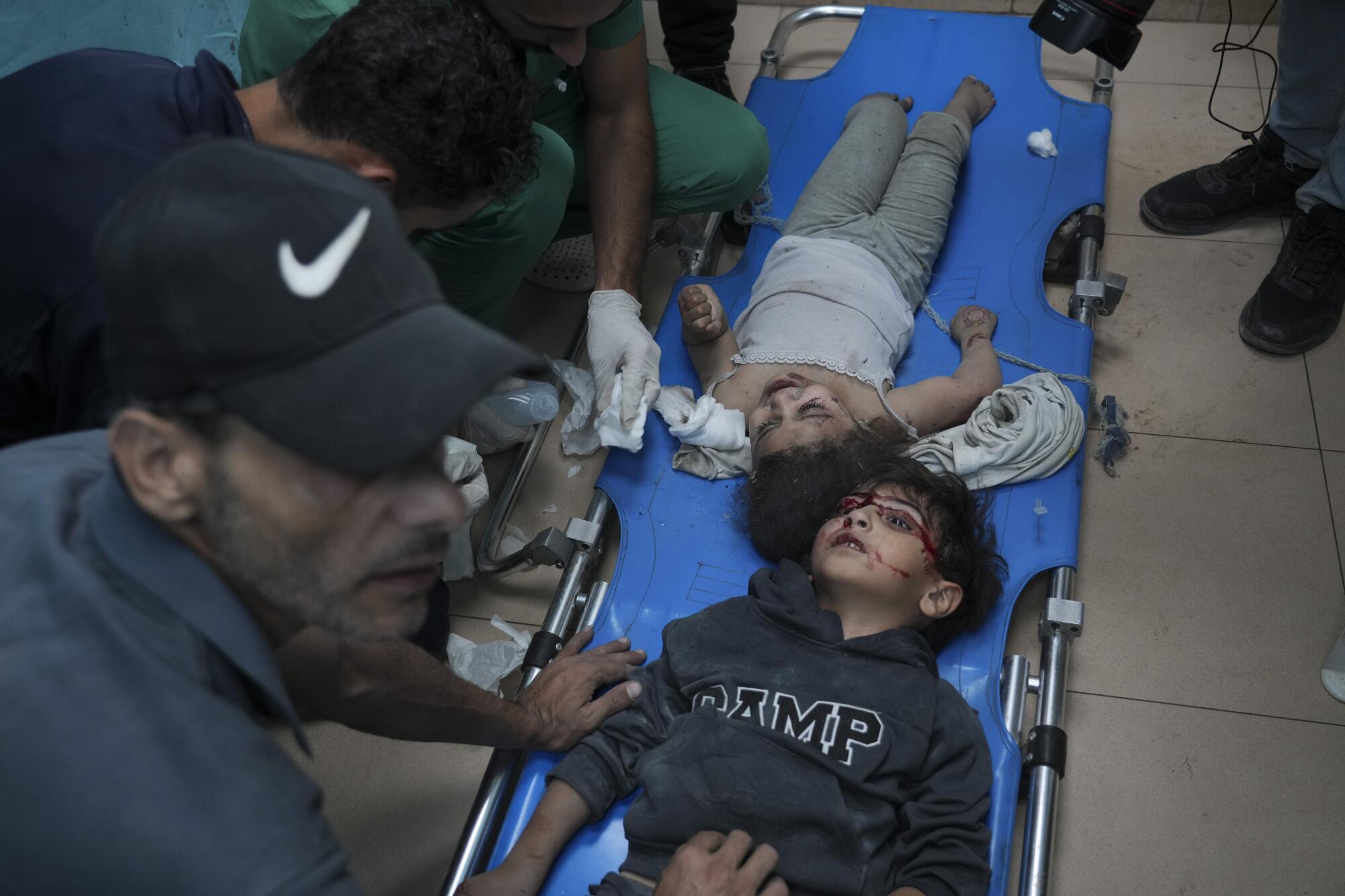 Two young children, one with blood on the face, lie on a blue stretcher as other people tend to them
