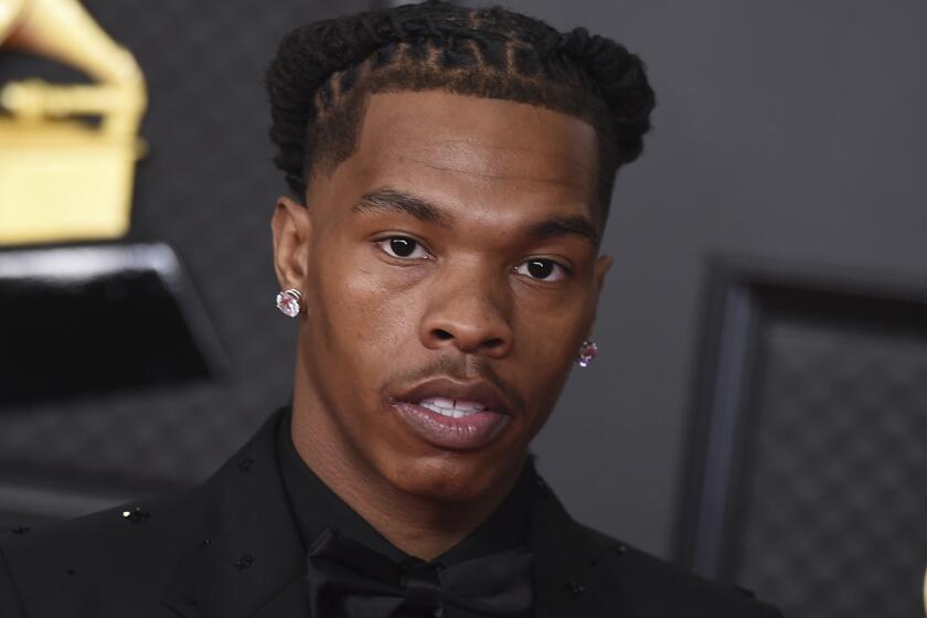 A Black man with a braided up-do and large stud earrings posing for pictures in a black suit